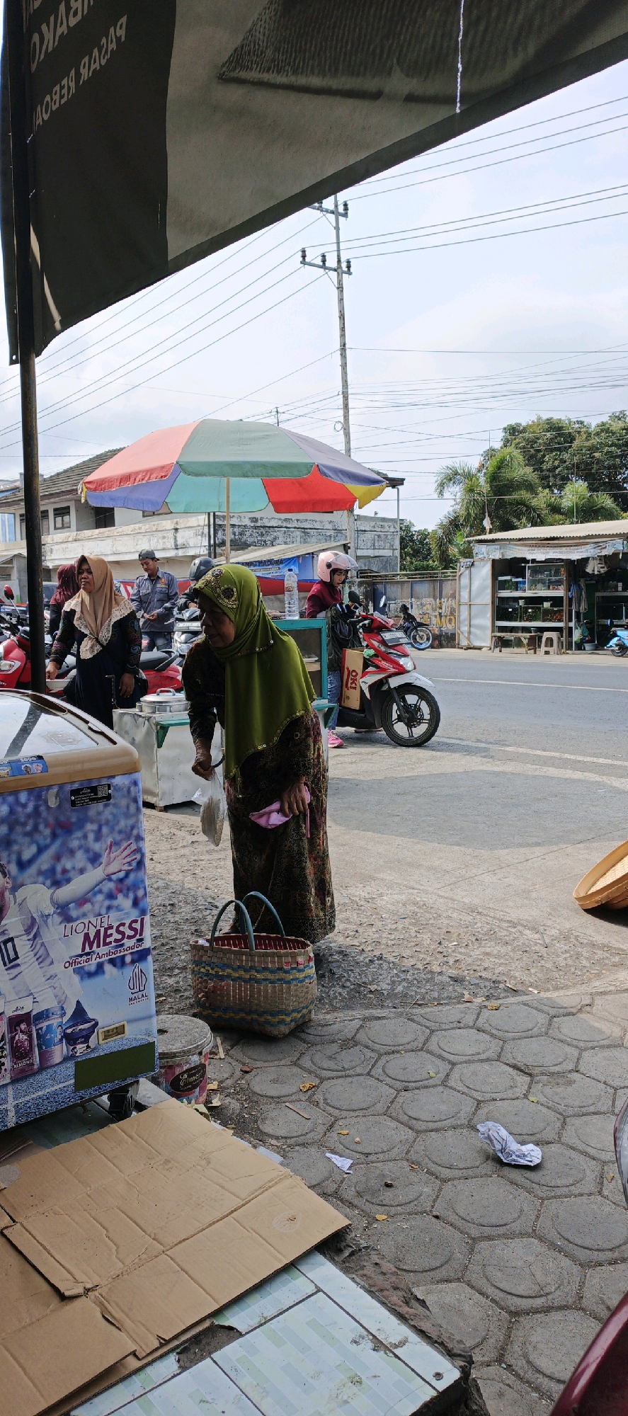 In Kasiyan, Indonesia, an outdoor market scene unfolds under a bright sky. A woman wearing a green hijab and a patterned dress stands near an ice cream freezer, holding a white plastic bag. Beside her rests a woven basket. In the background, a colorful umbrella provides shade for a small stall. Several motorcycles are parked around the market, and a woman wearing a pink helmet is seated on one. Buildings and utility poles are visible in the distance, suggesting a bustling urban environment. The ground is paved with hexagonal stones.