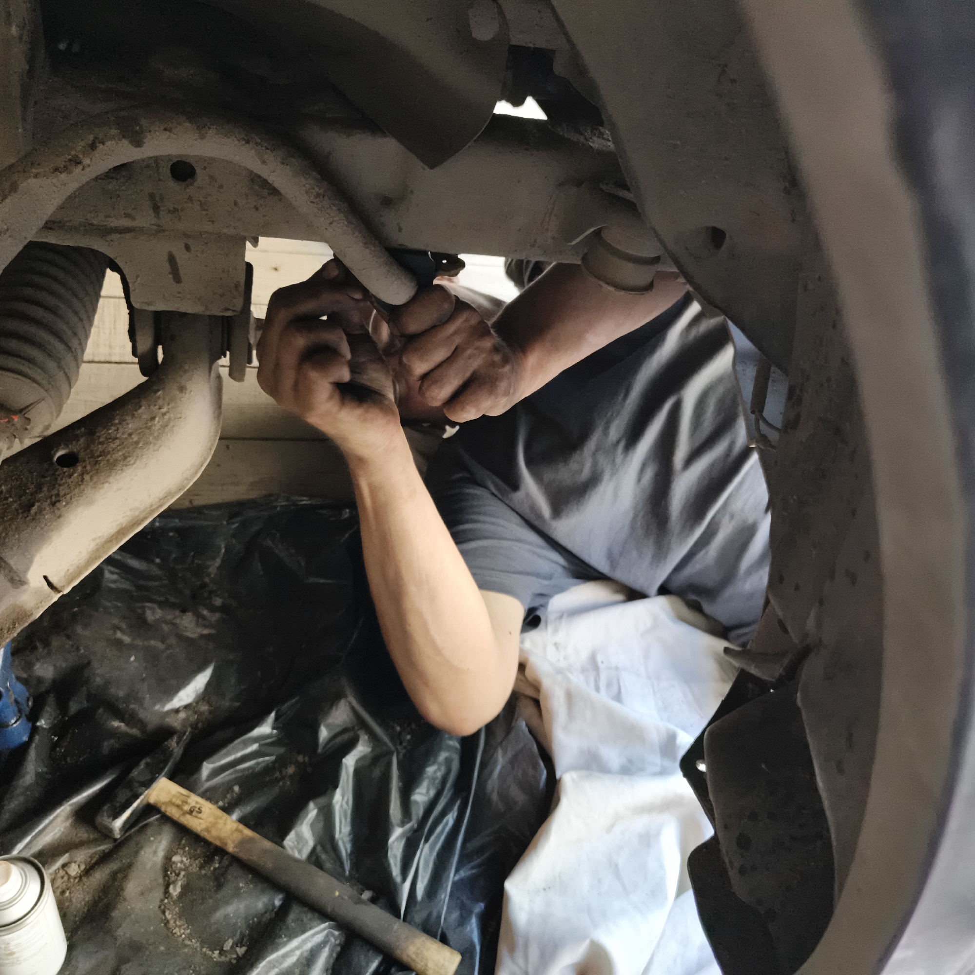 The image depicts a person working on the underside of a vehicle. The person is lying on their back on a black tarp, with their torso and arms visible. They are wearing a dark t-shirt and appear to be using tools to work on the car's suspension or chassis. Dirty hands suggest mechanical work. Various car parts, including suspension components and what appears to be a shock absorber boot, are visible. A hammer and a can of spray are on the tarp. The environment suggests an outdoor or garage setting, with wooden planks visible in the background. The image does not provide specific details to determine the exact location in Chaikovsky, Russia.
