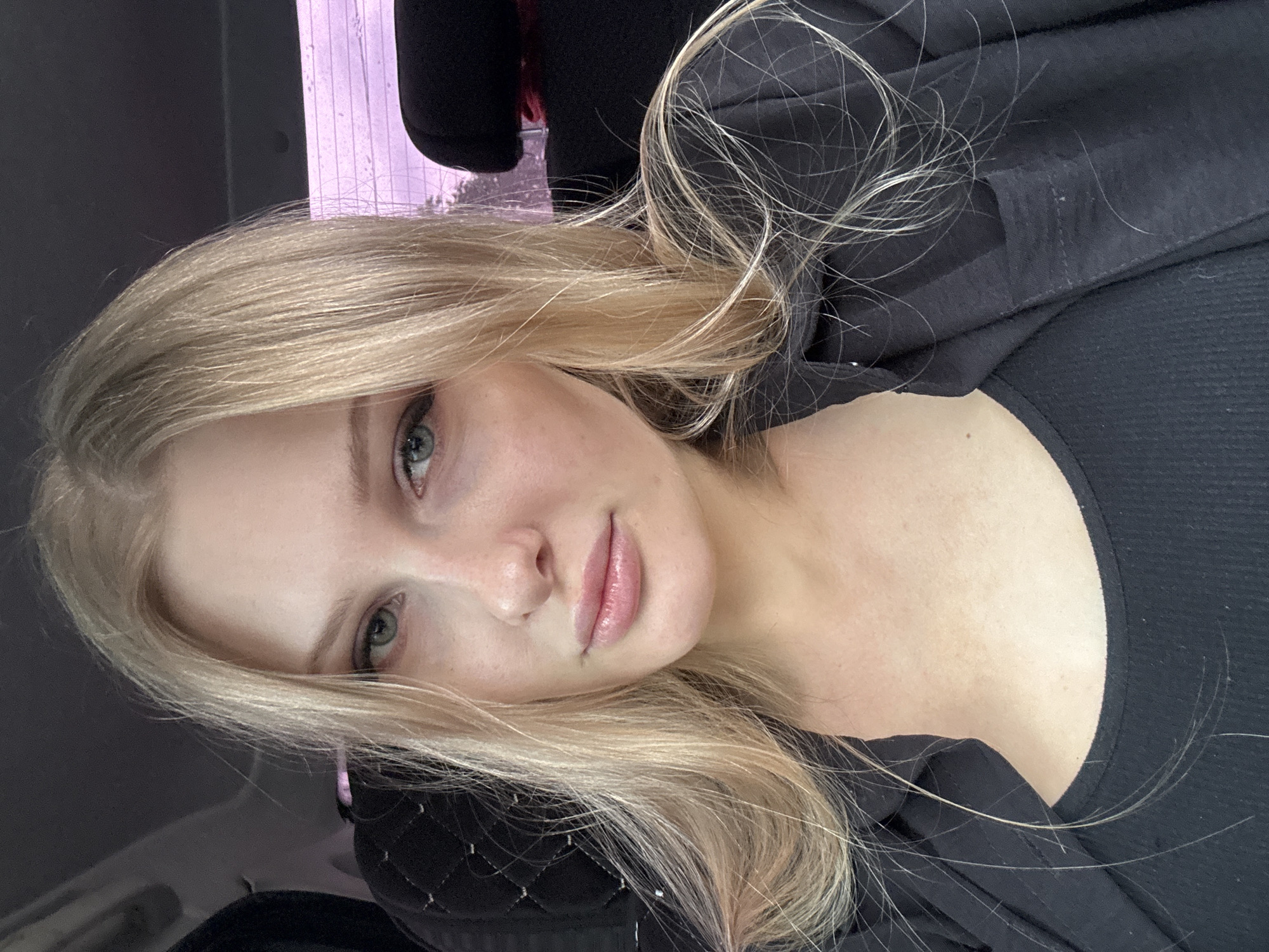 A young woman with blonde hair is posing for a selfie. She is wearing a black top and jacket. The background is blurry, but it appears to be the interior of a car or other vehicle. The location in the city of Perm, Russia, cannot be determined from the image.