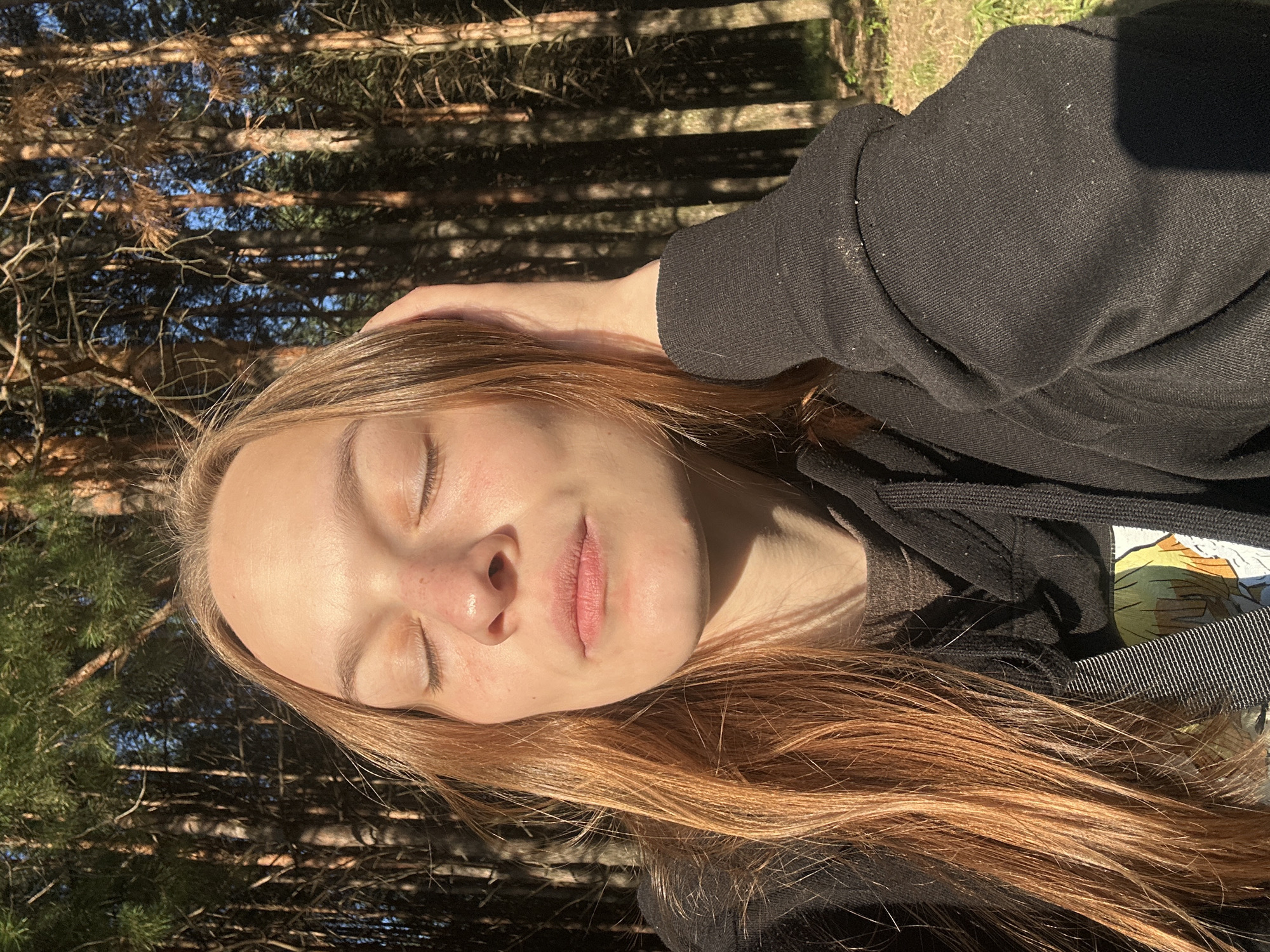 The image is a close-up, portrait-style selfie of a young person with long, auburn hair. They are wearing a black hoodie, and their eyes are closed, with a slight smile on their lips, suggesting a sense of peace or contentment. Sunlight streams down, casting dappled shadows on their face and highlighting the texture of their skin and hair. The background is a dense forest of pine trees, with sunlight filtering through the branches, indicating it is daytime. The overall mood is serene and natural, capturing a moment of quiet reflection outdoors. The image appears to be taken in Chaikovsky, Russia, given the context. No visible text is present in the image.