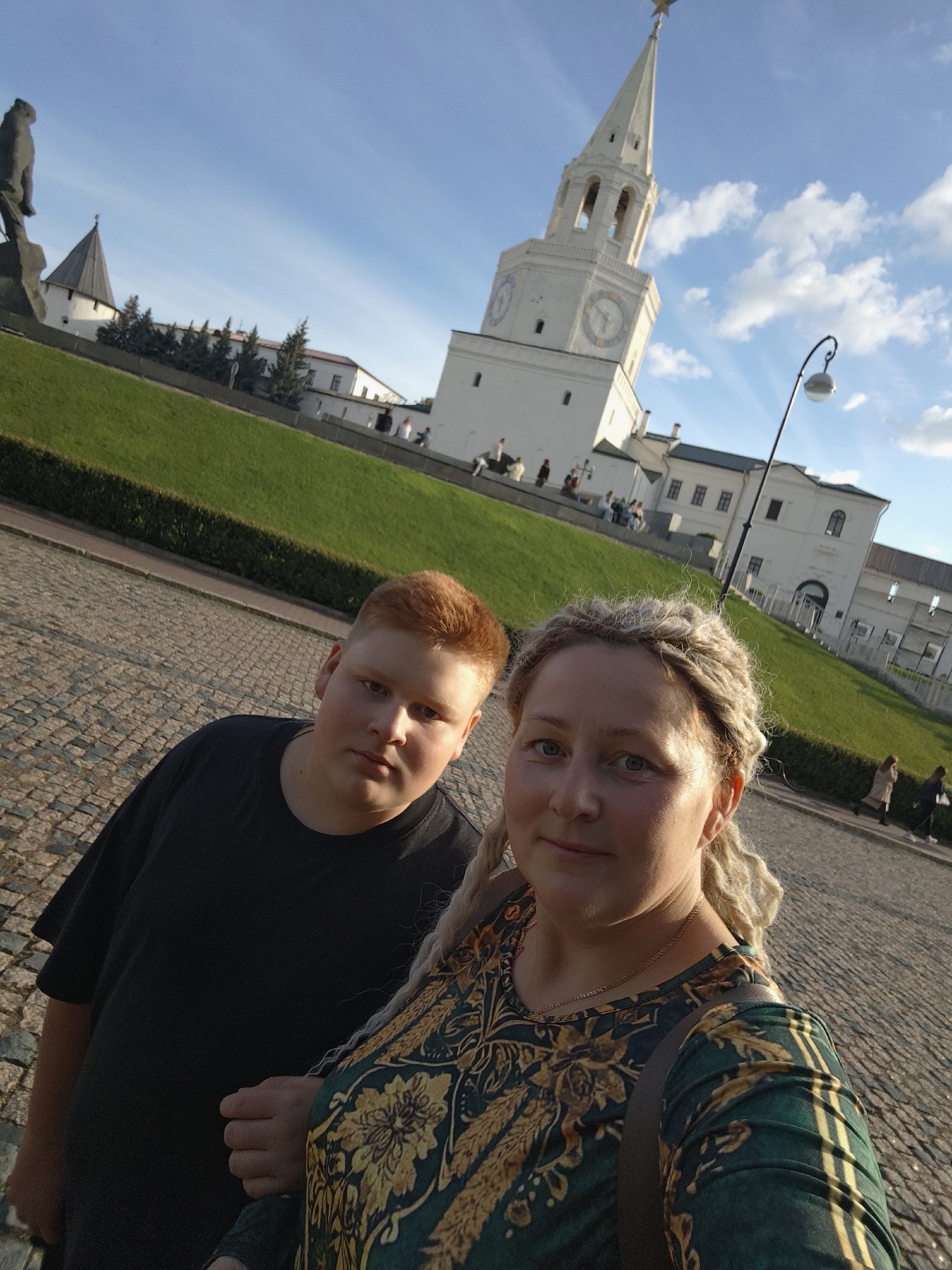 This media file is a selfie taken outdoors, featuring two people and a historic setting. In the foreground, a young person with reddish-blonde hair and a black t-shirt is positioned to the left, looking towards the camera with a neutral expression. To their right, a woman with blonde, braided hair and a patterned green and gold shirt is also looking at the camera, smiling softly. They appear to be standing on a cobblestone pathway.
Behind the subjects, a manicured green lawn slopes upwards towards a white building complex. Dominating the background is a tall, white tower with a pointed spire, featuring clock faces on its sides and a star at its apex. This tower is a prominent landmark, likely part of a fortress or historical site. Several smaller white buildings with red roofs are visible adjacent to the tower. The sky is blue with scattered white clouds, suggesting fair weather. The sunlight indicates it is likely late afternoon or early evening.
The overall scene suggests a tourist or sightseeing activity, with the subjects posing for a photo in front of a notable architectural landmark. The presence of other people in the background, some walking and some sitting, further supports this interpretation. The location is identified as Kazan, Russia, which aligns with the architectural style of the tower and buildings. No visible text is present in the image.