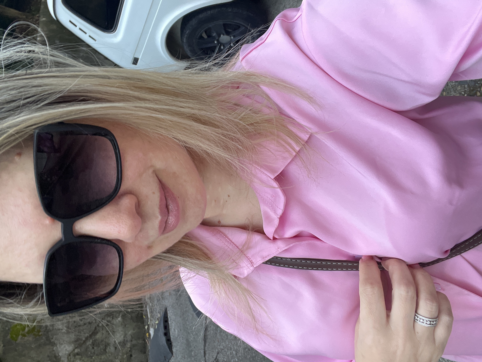A close-up selfie shows a light-skinned woman with blonde hair and pinkish lips, wearing large black sunglasses and a satin pink button-up shirt. Her left hand, adorned with a diamond ring, is resting on the shirt. A dark brown strap with white stitching is visible across her chest. In the background, a white car with black wheels is parked, and a rough concrete wall is to the left. The ground appears to be asphalt or pavement. The overall impression is a candid snapshot taken outdoors in an urban setting.