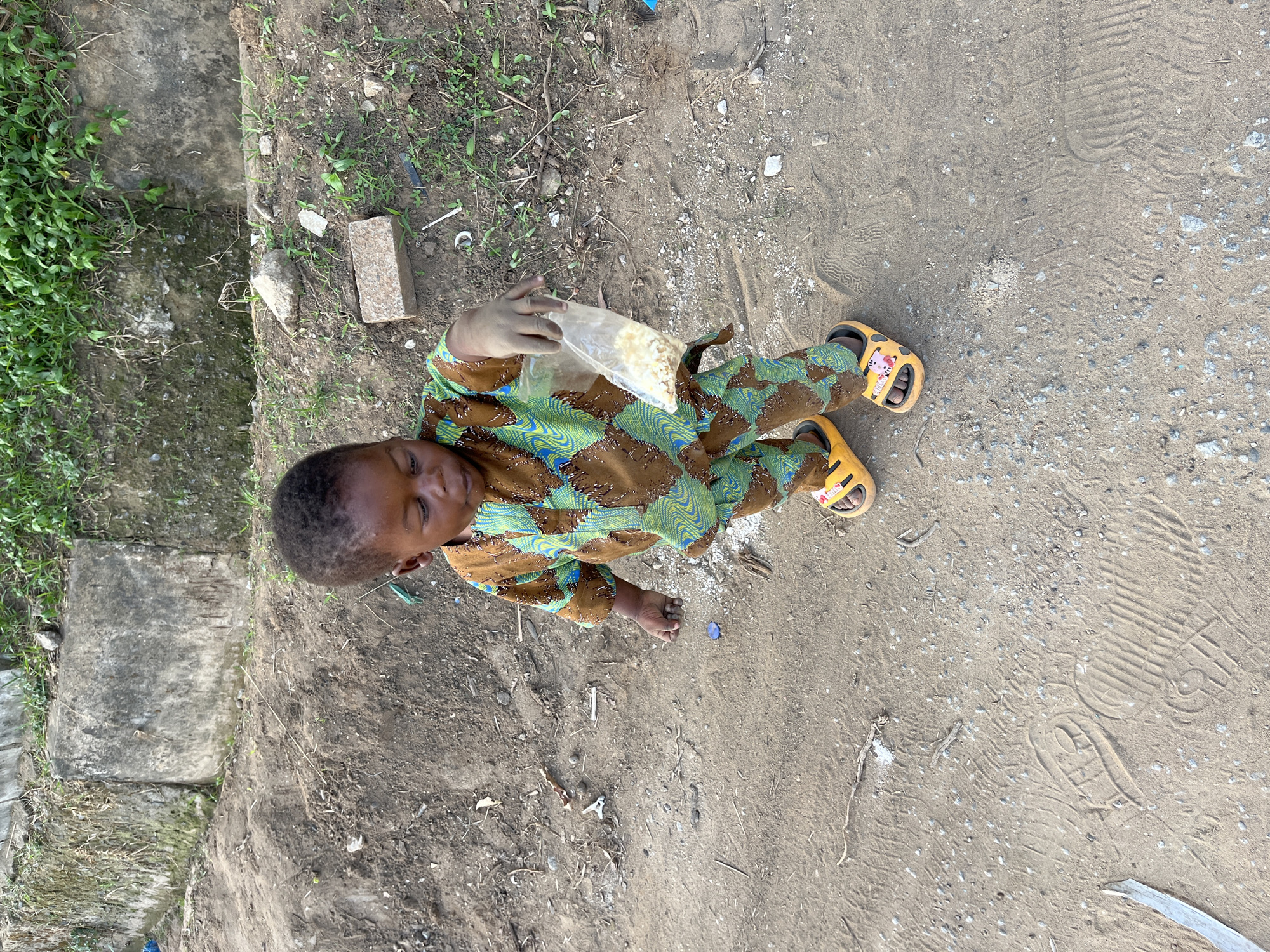A child in a patterned top and yellow sandals sits on dirt and holds a bag of food. The setting appears to be outdoors, possibly in a less developed area of Abuja, Nigeria, given the presence of dirt, debris, and simple construction.