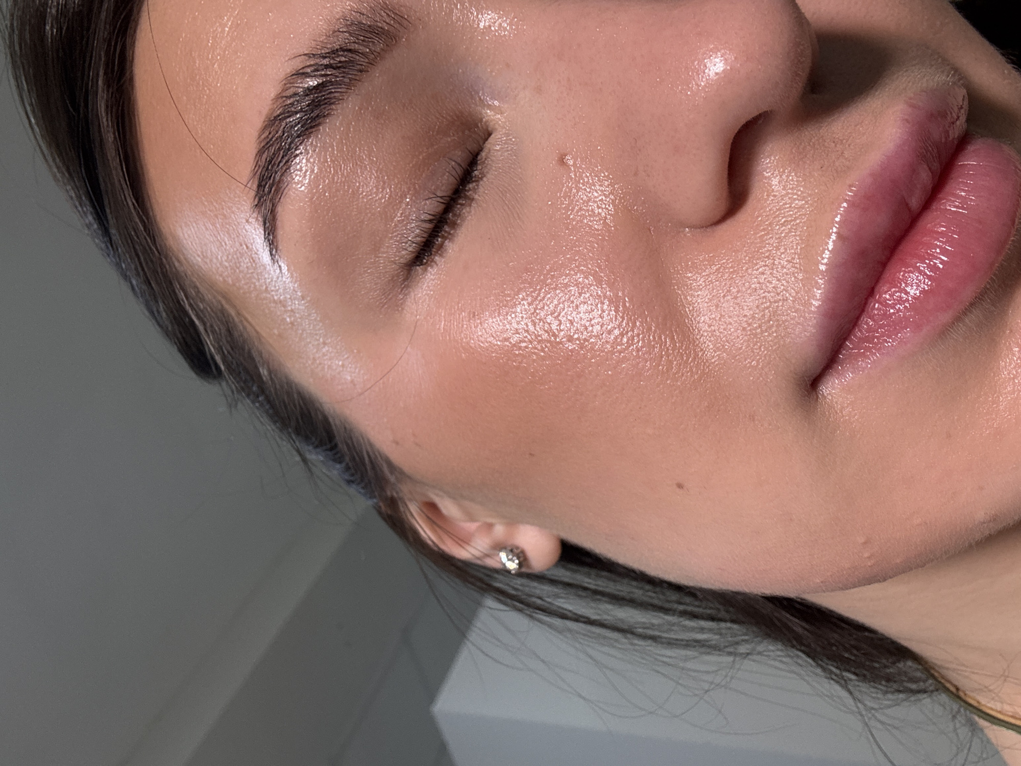 A close-up image shows a portion of a woman's face, specifically the area including her forehead, closed left eye, nose, cheek, and mouth. The skin is smooth and has a dewy or shiny appearance. The lips are full and glossy. The eyebrows are dark and well-groomed. A small earring is visible in the earlobe. The image's background is out of focus and appears to be a neutral gray or white. There is no information to suggest this image was taken in Chaikovsky, Russia.