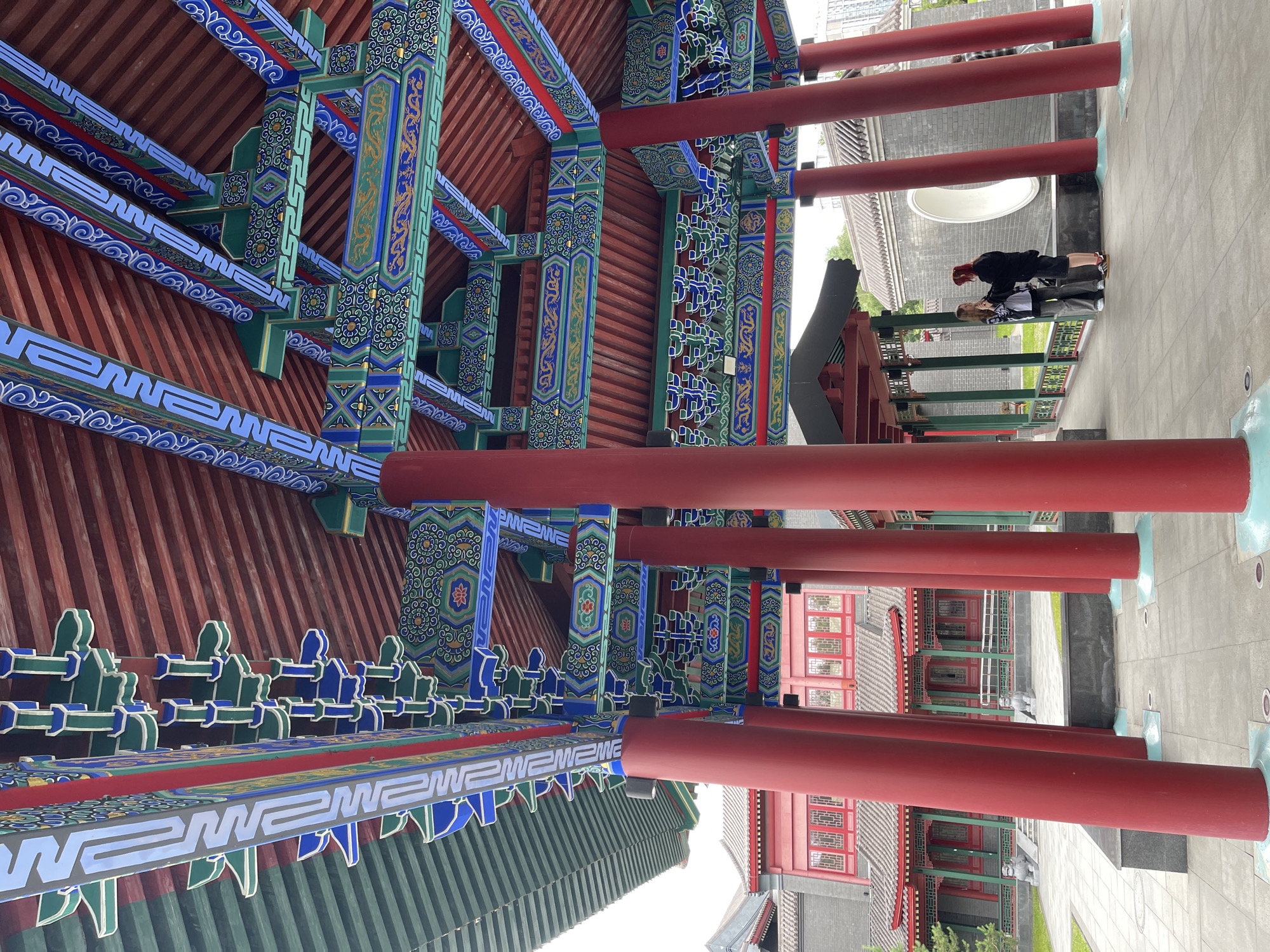 The image displays a traditional East Asian architectural style, characterized by red pillars, intricate painted beams, and red tiled roofs. In the foreground, the ornate wooden beams are richly decorated with patterns in blue, green, and gold, featuring motifs that appear to be dragons or other mythical creatures. These beams are part of a covered walkway or pavilion.
In the mid-ground, two individuals are visible, standing close together and appearing to interact. One person, wearing a black jacket and grey pants, is leaning slightly towards the other, who is dressed in a patterned shirt and grey pants. They seem to be engaged in a conversation or sharing a moment. Beyond them, a courtyard area is visible with more traditional buildings featuring similar architectural elements. Stone statues, possibly of lions or guardian figures, are positioned near the buildings.
The setting appears to be a cultural or historical site, possibly a temple complex or a heritage area. The lighting suggests it is daytime, and the weather seems pleasant, with bright but diffused light. The overall impression is one of cultural richness and a serene atmosphere. There is no visible text in the image.