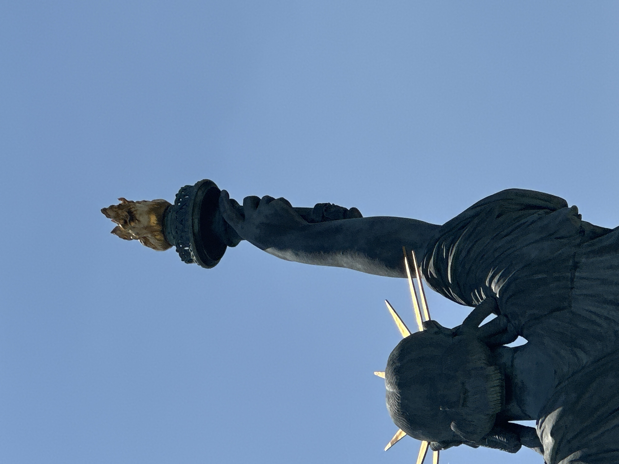 The image shows a close-up, low-angle view of the torch held aloft by the Statue of Liberty on the Île aux Cygnes in Paris, France. The sky is a clear, pale blue. The torch itself is made of gilded metal and appears to be lit with a bright, warm light. The statue's dark, textured arm and hand are visible gripping the torch. The back of the statue's head, with its characteristic crown of rays, is also visible, also rendered in dark, textured material. The image is cropped to focus on the upper portion of the statue and the torch against the expansive sky.