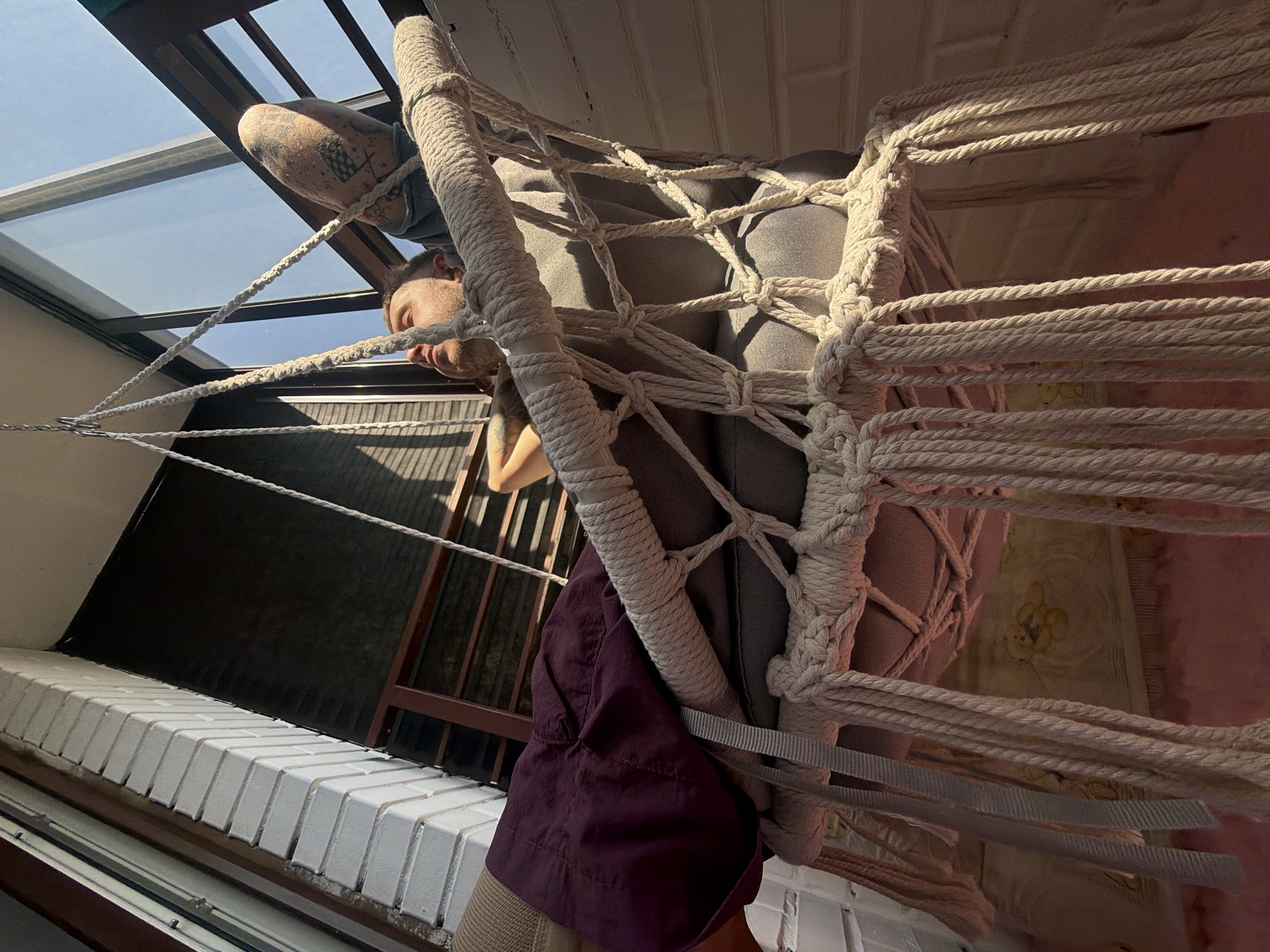 A man with tattoos on his arm is seated in a hanging macrame chair inside a room with a skylight. He is wearing a dark shirt, light pants, and a burgundy jacket. The room has a white brick windowsill, and a staircase is visible through the ropes of the chair. The city of Saint Petersburg, Russia is not evident in this image.