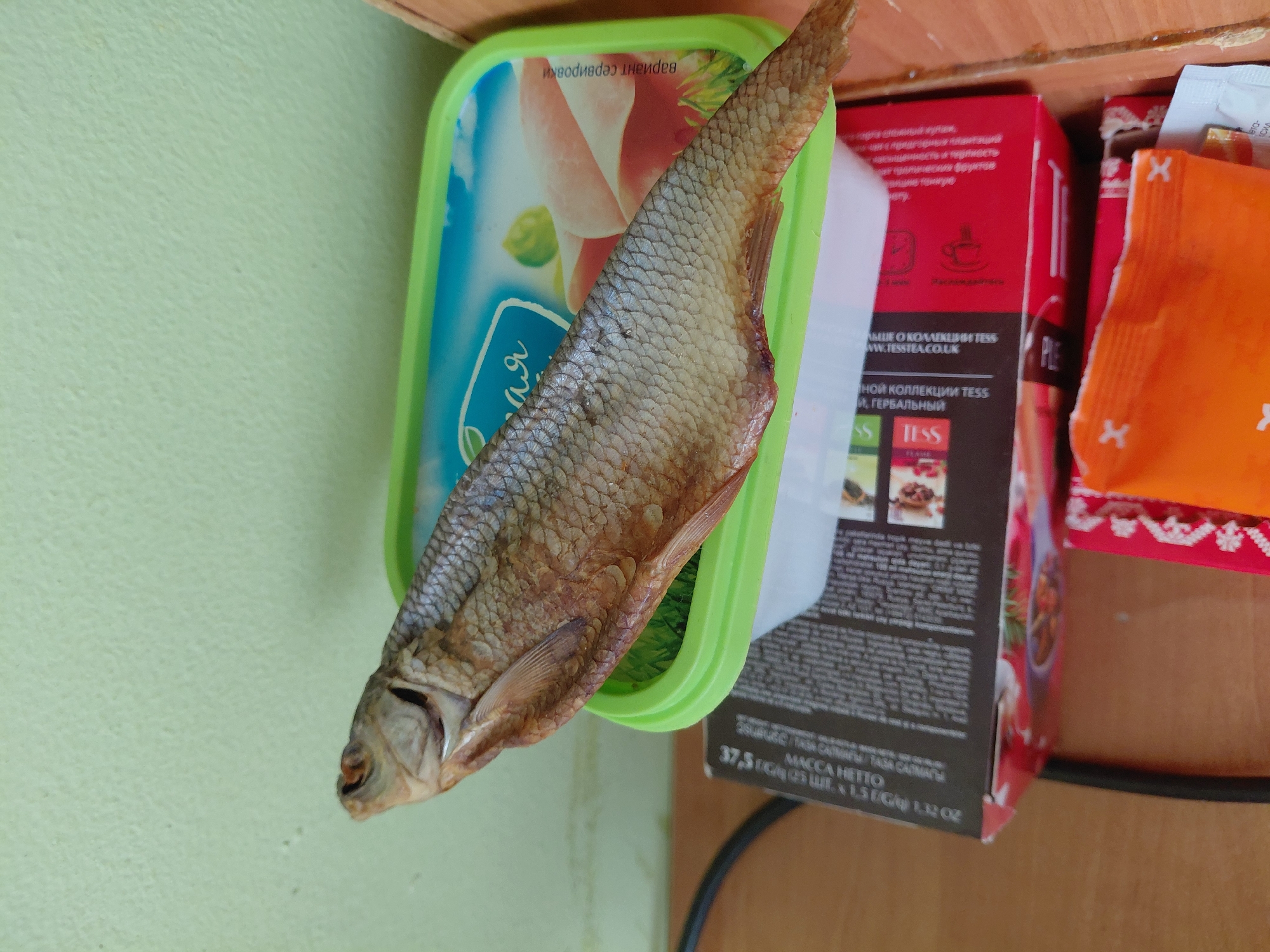 The image displays a close-up, top-down view of a dried fish resting on a light green-rimmed plastic container, with various food packages in the background, set against a light green wall and a wooden surface in what appears to be an indoor setting in Nadym, Russia.
The main subject is a whole, dried fish, possibly a vobla, positioned diagonally. Its textured, scaly body, head with a visible eye, and fin details are clearly seen, indicative of a dried seafood product.
The fish is placed on a light green plastic container featuring a translucent blue lid. Russian text "Для" (For) is visible on the lid, along with "вариант сервировки" (serving option/variant) printed upside down. An indistinct image of sliced food, possibly cured meat or fish, is visible on the lid's graphic.
Behind this container are several packaged goods. A prominent red box for TESS tea is visible, displaying information about its collection in both Russian and English, including "БОЛЬШЕ О КОЛЛЕКЦИИ TESS WWW.TESSTEA.CO.UK" (More about the TESS collection) and "ИНОЙ КОЛЛЕКЦИИ TESS И, ГЕРБАЛЬНЫЙ" (Other collection TESS, herbal). The net weight is stated as "MACCA HETTO 37,5 Г / G / ШТ. X 1.5 Г / G / 1.32 OZ". To the right of the tea box, a partially visible orange packet with an "X" logo is present, likely another consumer product.
No specific activity or event is taking place; the items appear to be stored or temporarily placed. The lighting is even, suggesting an indoor environment. No people are visible, and no emotions are conveyed.