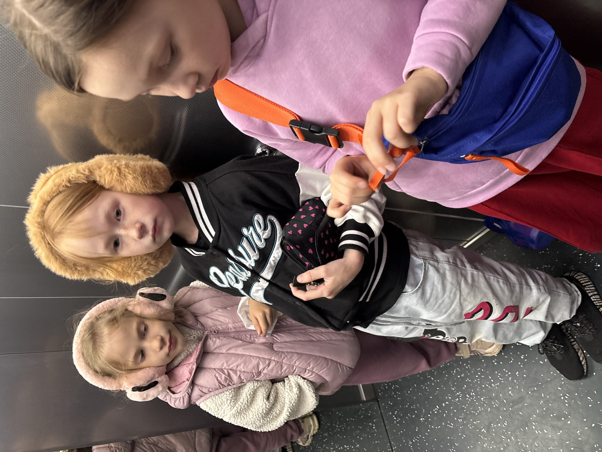 Three young girls are standing in an elevator. One girl is wearing a pink hoodie and a blue and orange bag. Another girl is wearing a black jacket, light-colored pants, and earmuffs. The third girl is wearing a pink jacket, a white fleece, and earmuffs. The elevator's interior is gray with a speckled floor. There is no visible information indicating the location of Izhevsk, Russia.