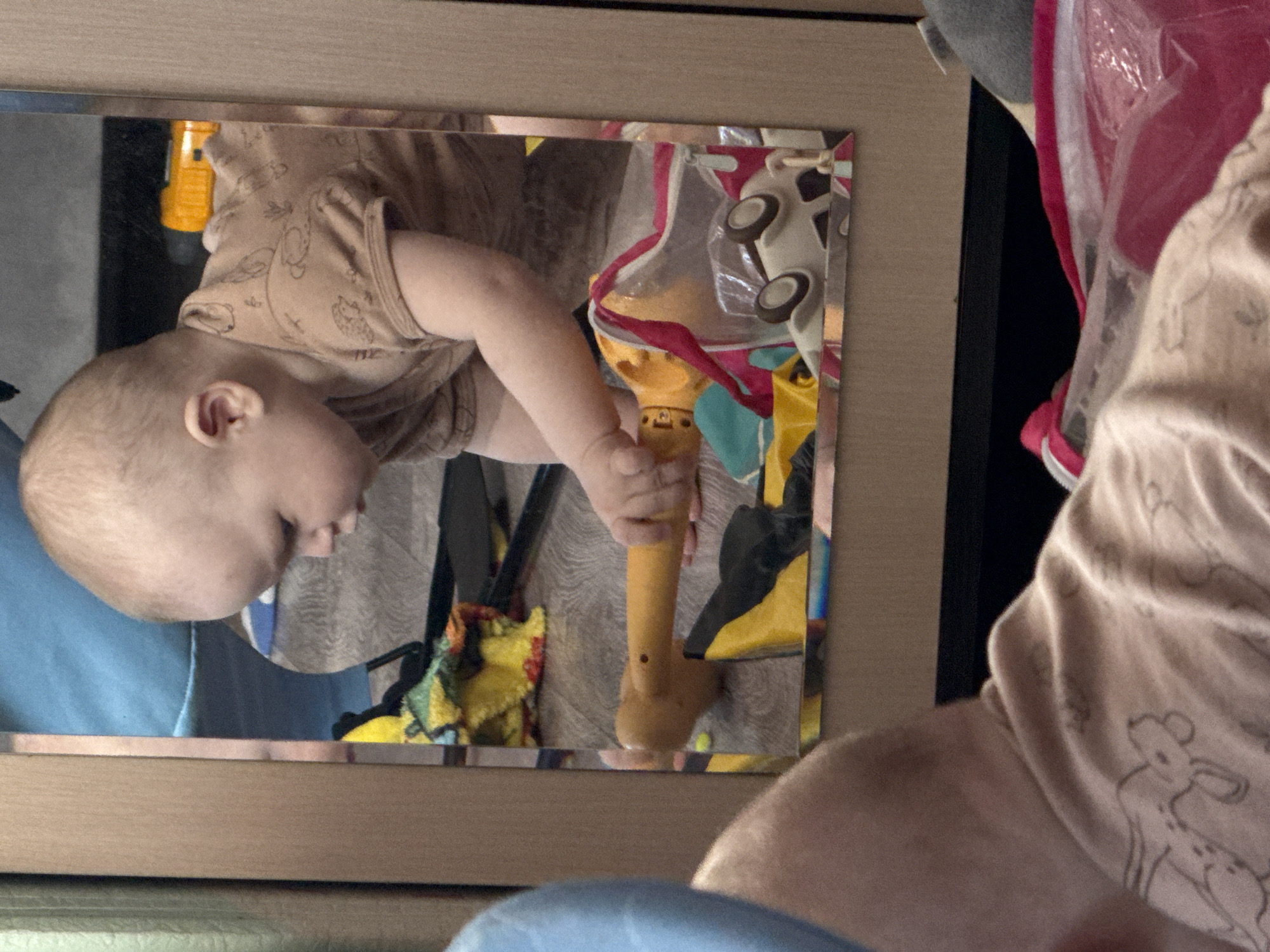 A baby wearing a brown short-sleeved shirt is reflected in a mirror, holding a yellow toy. Other toys and objects are visible in the mirror's reflection. The baby is positioned on the left side of the frame, with the mirror filling the center. Parts of another person are visible on the right side of the frame, also wearing a brown shirt. The environment appears to be indoors. Location Кипчак, Russia is not discernible.