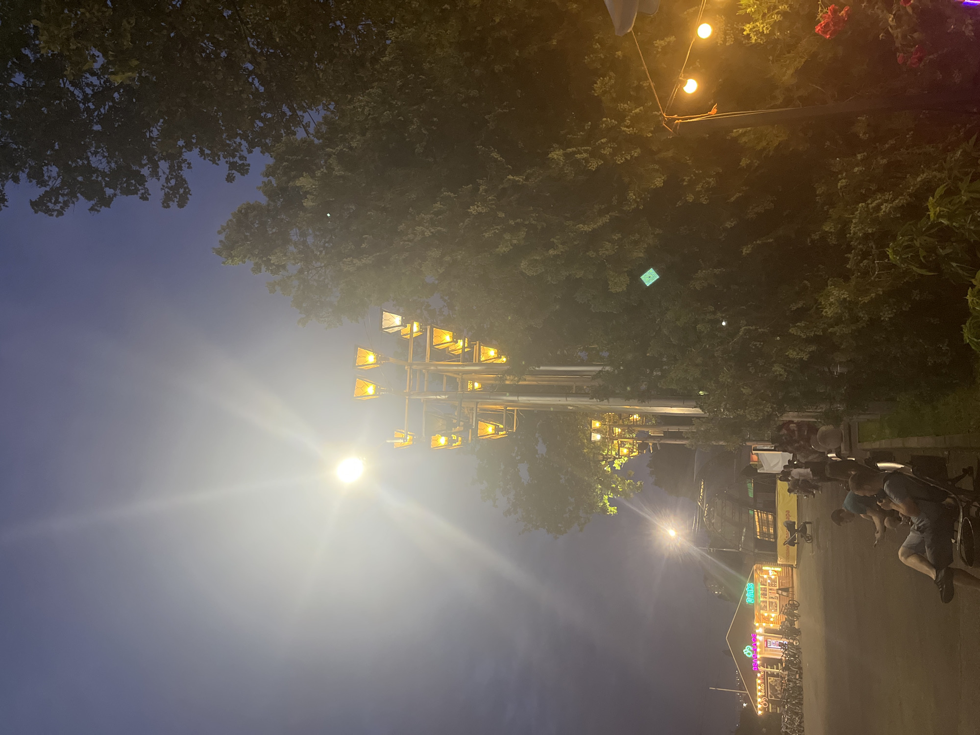 The image depicts an outdoor evening scene at a cafe in Krasnodar, Russia, identified by the signage which reads "ВЕЛОКАФЕ" (Velocafe). The cafe appears to be a popular gathering spot, with several bicycles parked outside. A prominent street lamp with multiple yellow lights illuminates the scene, casting a bright glow. In the foreground, a few people are seated on benches, with one individual looking at their phone. The background shows the cafe structure, some greenery, and a dimly lit sky. The overall atmosphere suggests a casual and relaxed evening at an urban cafe.