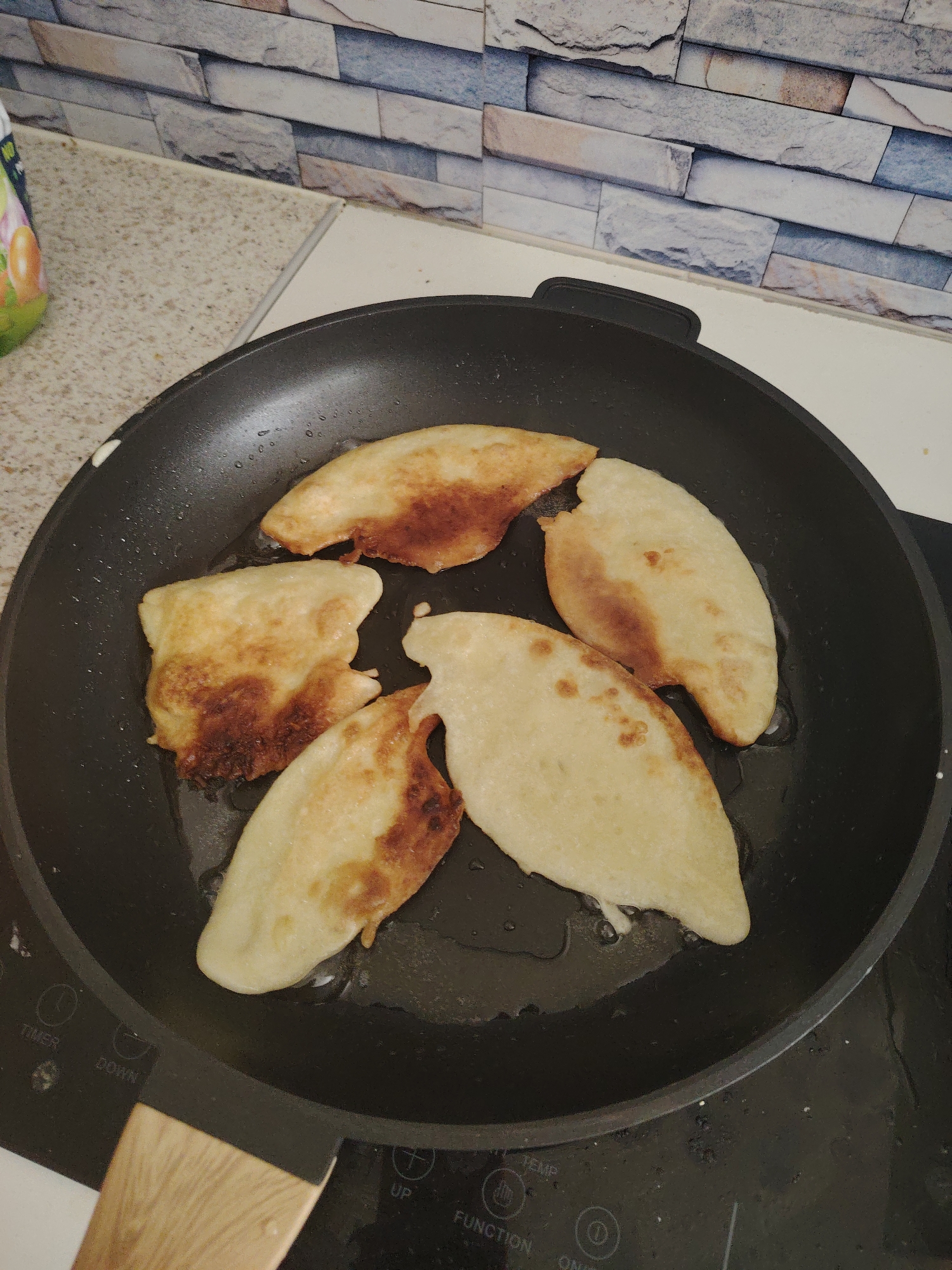 The image displays a close-up shot of a black frying pan on a stovetop, containing five pieces of dough actively cooking in oil. The dough items, which resemble dumplings or flatbreads, are partially browned and golden, indicating they are in the process of being fried. Some areas show darker, crispier browning, while others remain a lighter, un-fried color.
The frying pan has a black non-stick surface and a light-colored wooden handle, part of which is visible in the lower left corner. It rests on a sleek, black stovetop, likely an induction or electric hob, where faint white text and symbols are visible, including "TIMER," "DOWN," "UP," "FUNCTION," "ON/OFF," and "TEMP" controls.
The setting is a modern kitchen. To the left of the pan, a light speckled countertop is visible, and further to the left, a colorful bottle or container with indistinct labels can be seen. The background features a kitchen backsplash or wall covered with rectangular tiles in various shades of grey, beige, and white, arranged in a brick-like pattern.
The image captures an everyday cooking activity, focusing on the food preparation. There are no people visible, and no specific time of day, weather, or emotions can be discerned. Based on the provided context, this kitchen scene is located in Huntingdonshire, United Kingdom.