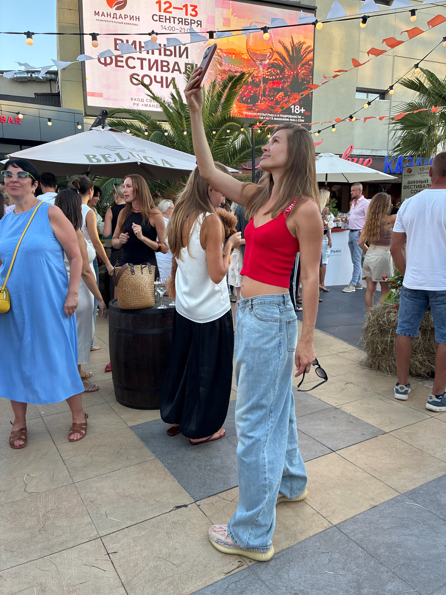 A medium shot captures a group of people in an outdoor urban setting, possibly a festival or market, in Sochi, Russia. In the foreground, a woman in a red crop top and light blue wide-leg jeans holds up a smartphone, taking a selfie. She is looking up and to her right. To her left, a woman in a light blue dress with a yellow purse walks away from the camera, her back mostly to the viewer. She wears brown sandals. Behind her, other individuals are visible, some interacting, some looking towards the large screen in the background.
The large screen displays text in Russian, indicating an event is taking place. The text reads "МАНДАРИН" (Mandarin), "12-13 СЕНТЯБРЯ" (September 12-13), and "ВИННЫЙ ФЕСТИВАЛЬ СОЧИ" (Wine Festival Sochi). This strongly suggests the location is the Mandarin Mall in Sochi, Russia, and the event is a wine festival occurring on September 12th and 13th. The screen also shows an image of a wine glass and palm trees.
Various elements of the environment are visible, including white umbrellas, string lights with decorative flags, a wooden barrel, a pile of hay, and paved ground with a patterned tile design. The lighting suggests it is daytime, possibly late afternoon or early evening. The overall atmosphere appears festive and crowded.