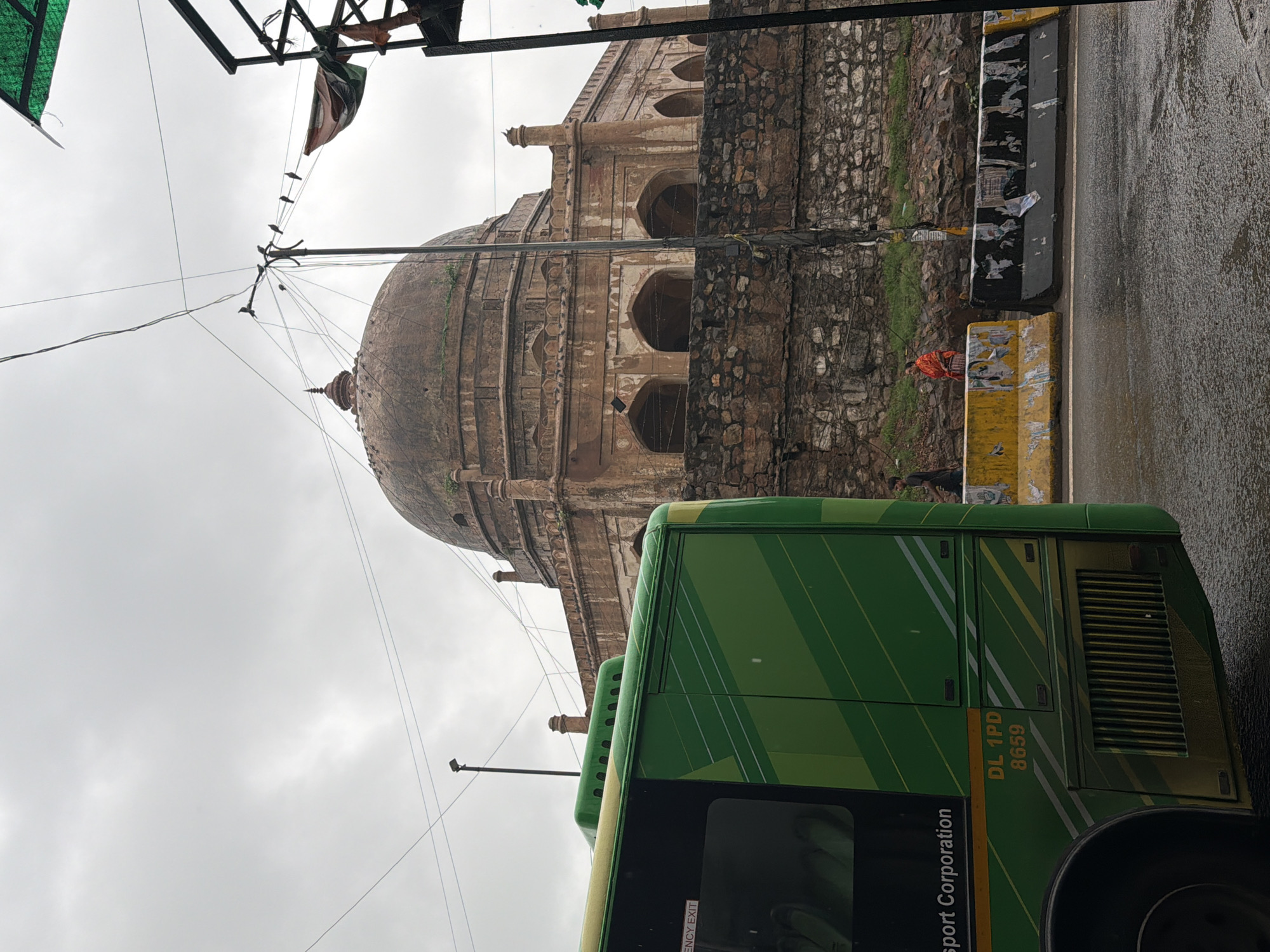 The image captures a segment of the historical Jamali Kamali Mosque and Tomb complex in Mehrauli, Delhi, India. In the foreground, the rear section of a green public transport bus is visible on the left, displaying the registration number "DL 1PD 8659". The bus has a distinctive green and yellow striped design. To the right of the bus, a stone wall demarcates the complex, and beyond it, the domed structure of the tomb, constructed from reddish-brown stone, dominates the background. Several arches are visible on the lower facade of the tomb structure. The sky is overcast, suggesting a cloudy day. In the middle ground, concrete barriers, one yellow and one black, line the road. Two individuals are standing near these barriers. The black barrier is partially covered with torn posters. A man in a dark shirt and trousers stands on the left, and a woman in an orange outfit stands to his right. The road surface appears wet, indicating recent rain. Overhead, electrical wires crisscross the sky, and a lamppost is visible. Birds are perched on the wires.