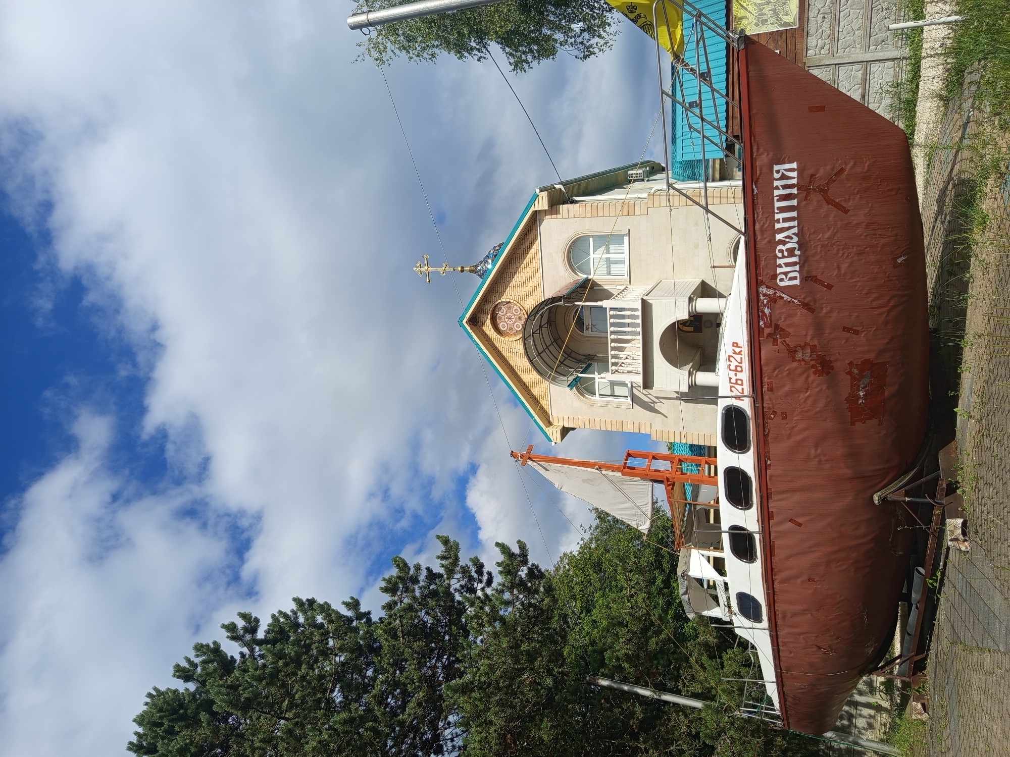 In the city of Giaginskaya, Russia, an old, partially weathered brown and white sailboat is displayed on a stand on a paved area. The sailboat is positioned in front of a tan, two-story building that appears to be a church, complete with a cross on its roof. The building features arched windows and a balcony. To the left of the sailboat, a large, green pine tree stands tall against a cloudy blue sky. The scene captures a static display with no people or activity evident.