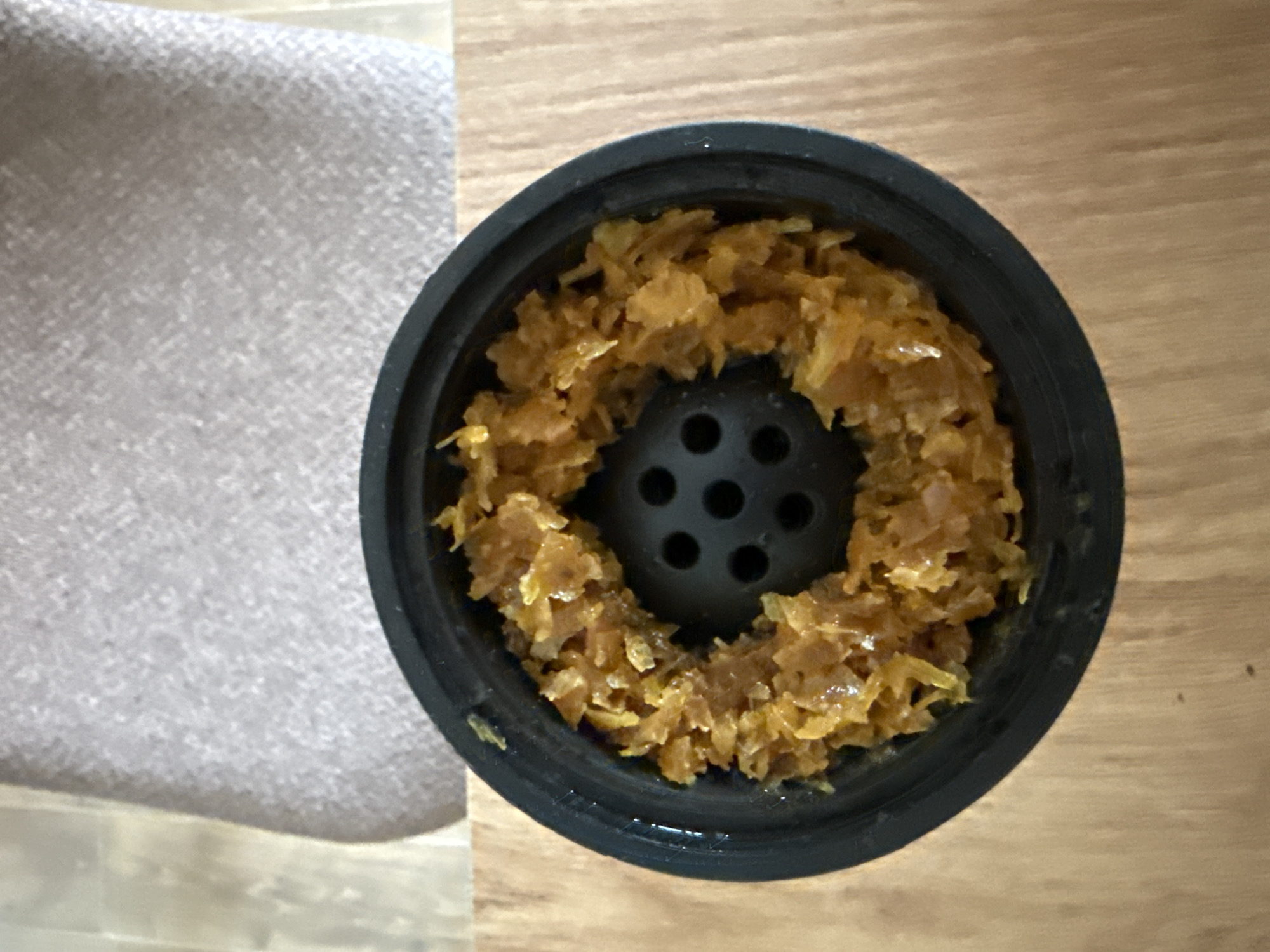 The image displays a top-down view of a black, circular hookah phunnel bowl filled with shisha tobacco. The tobacco is a moist, shredded substance with a prominent brownish-orange color and a glossy sheen, indicating the presence of molasses or glycerin. It is meticulously packed in a ring around the central, raised, and perforated spire of the bowl.
The black bowl itself appears clean and has a modern design. It rests on a light-colored wooden surface, likely a table, which is visible on the right side of the frame. To the left, a soft, light grey textured fabric, possibly upholstery from a couch or chair, occupies part of the background. This arrangement suggests an indoor setting, likely a casual lounge or living space, in Varna, Bulgaria.
The primary activity depicted is the preparation for a hookah smoking session, with the shisha tobacco loaded into the bowl. No people are visible, and there is no discernible text within the image. The lighting is consistent with an indoor environment, making it impossible to determine the time of day or weather conditions. The scene evokes a sense of readiness for a relaxed, social gathering.