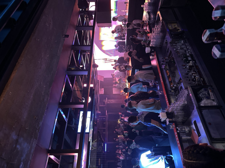 The image depicts the interior of Club Yakata, a nightclub located in Sofia, Bulgaria. The scene is dimly lit with prominent purple and blue ambient lighting.
In the foreground, a black bar counter extends across the bottom right, equipped with stainless steel workstations, multiple clear glassware stacks, ice buckets, and numerous bottles of spirits and mixers arranged on shelves. A napkin dispenser and fruit garnishes are also visible on the counter. A person with braided hair, facing away from the camera, is partially visible in the bottom left, positioned at the bar.
The midground features a large crowd of approximately 50-70 individuals, primarily young adults, standing and socializing. They are distributed throughout the club space, with a significant number gathered along the bar. Interactions appear social, with people conversing and holding drinks. To the right of the crowd, an elevated DJ booth is present with visible DJ equipment, including mixers and turntables. Two individuals are positioned behind the DJ console.
The background reveals large illuminated screens or panels displaying abstract graphics, one of which prominently features a large white "W" shape. Further left in the background, another illuminated sign on the wall spells out "YAK". The club's ceiling features exposed industrial-style structures, including metal trusses and beams, with embedded lighting fixtures casting colored light. No overt actions such as dancing are clearly discernible, but the overall atmosphere suggests a social gathering within a nightclub environment.