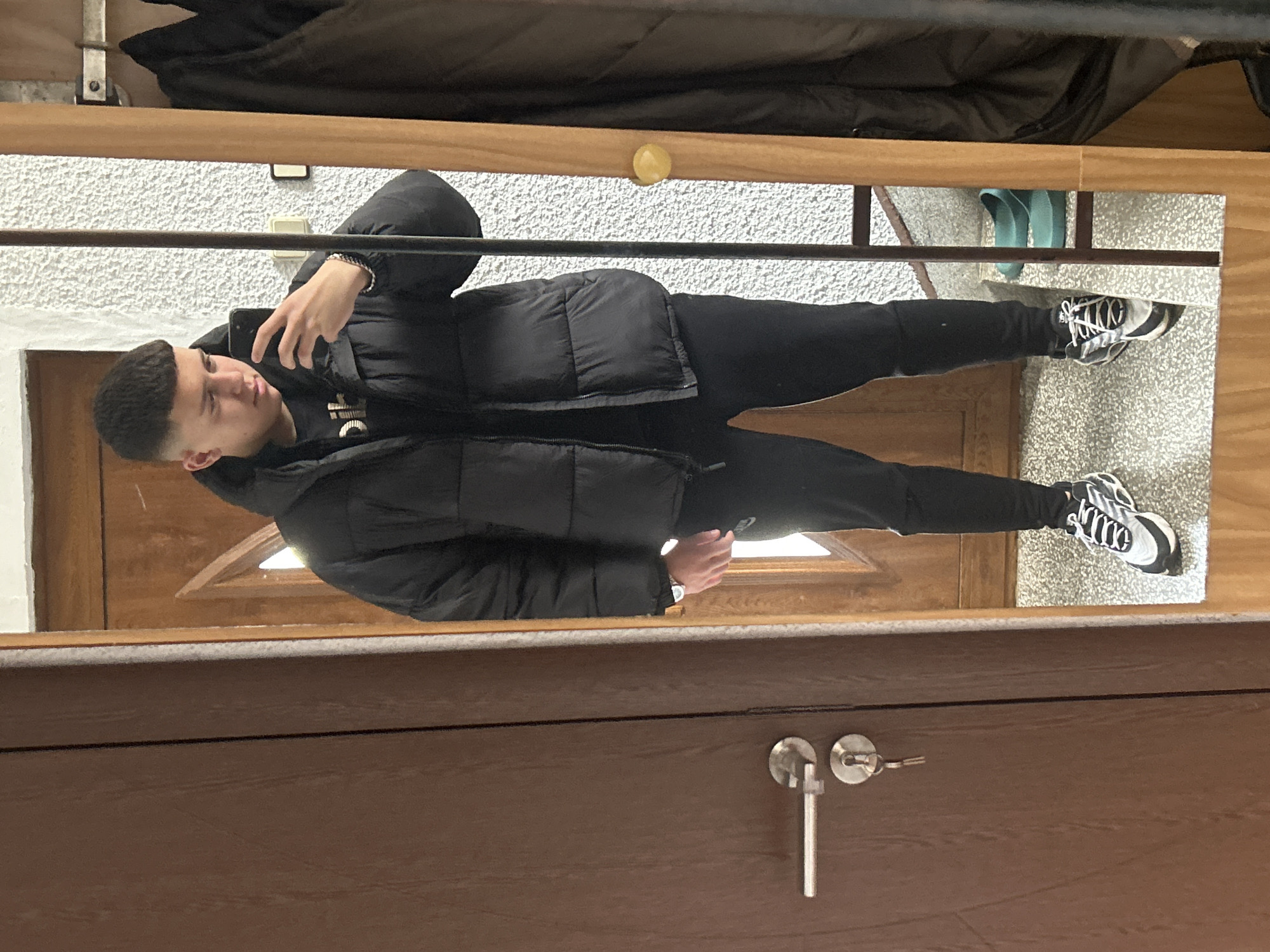 A young man is taking a selfie in a mirror. He is wearing a black puffer jacket, black sweatpants, and black and white sneakers. The reflection includes the interior of a doorway, including a dark-colored door with a silver handle and lock, a wall with a light-colored textured finish, and some personal items such as slippers. The location is possibly Zlatograd, Bulgaria.