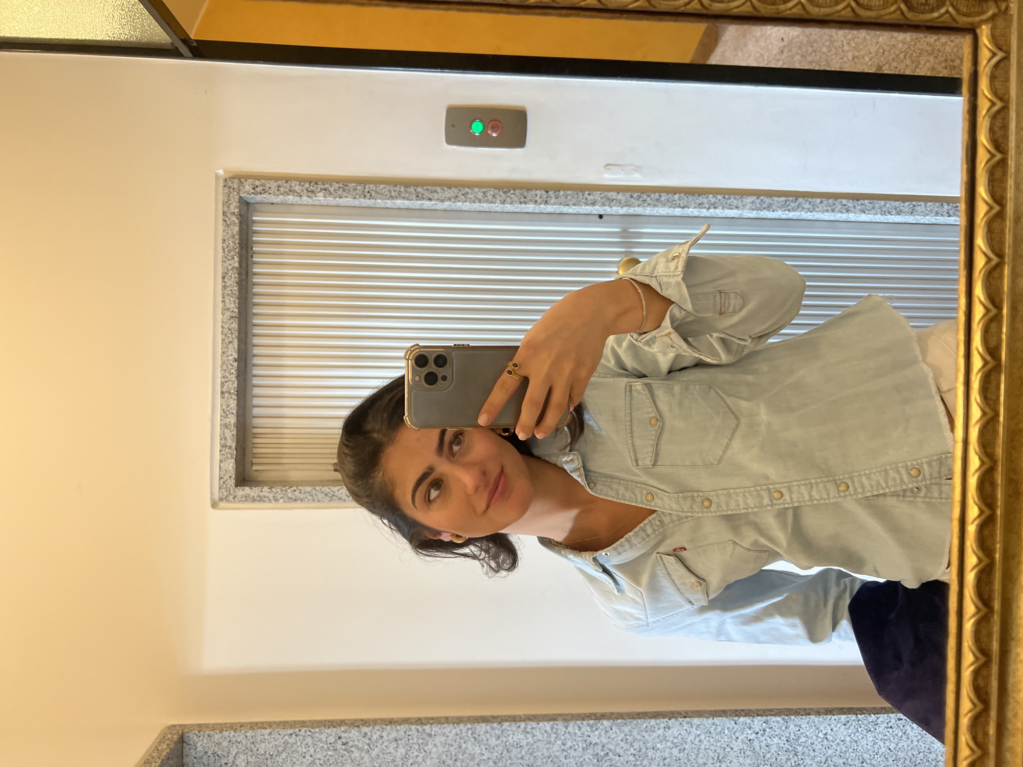 A young woman is captured in a mirror selfie, likely in an indoor setting in Milan, Italy. She is looking directly into the camera with a gentle smile. Her dark hair is pulled back, revealing a clean, expressive face. She is wearing a light blue denim button-up shirt. Her left hand, adorned with two rings and a delicate bracelet, holds a grey-cased smartphone to take the picture. A fine gold necklace and small gold hoop earrings are also visible.
The mirror she is using has a prominent, ornate gold frame. The background reflected in the mirror behind her primarily shows a light-colored wall. Above her reflection, an elevator control panel is visible on the wall, featuring two illuminated circular lights: a green one on the left and a red one on the right. Below this panel and to the side, a section of what appears to be an elevator door or a louvered vent with horizontal slats is reflected, framed by a speckled grey material. The lower part of the reflection also shows more of this speckled grey surface, possibly indicating flooring or a counter. The overall lighting is bright and even, consistent with an interior space.