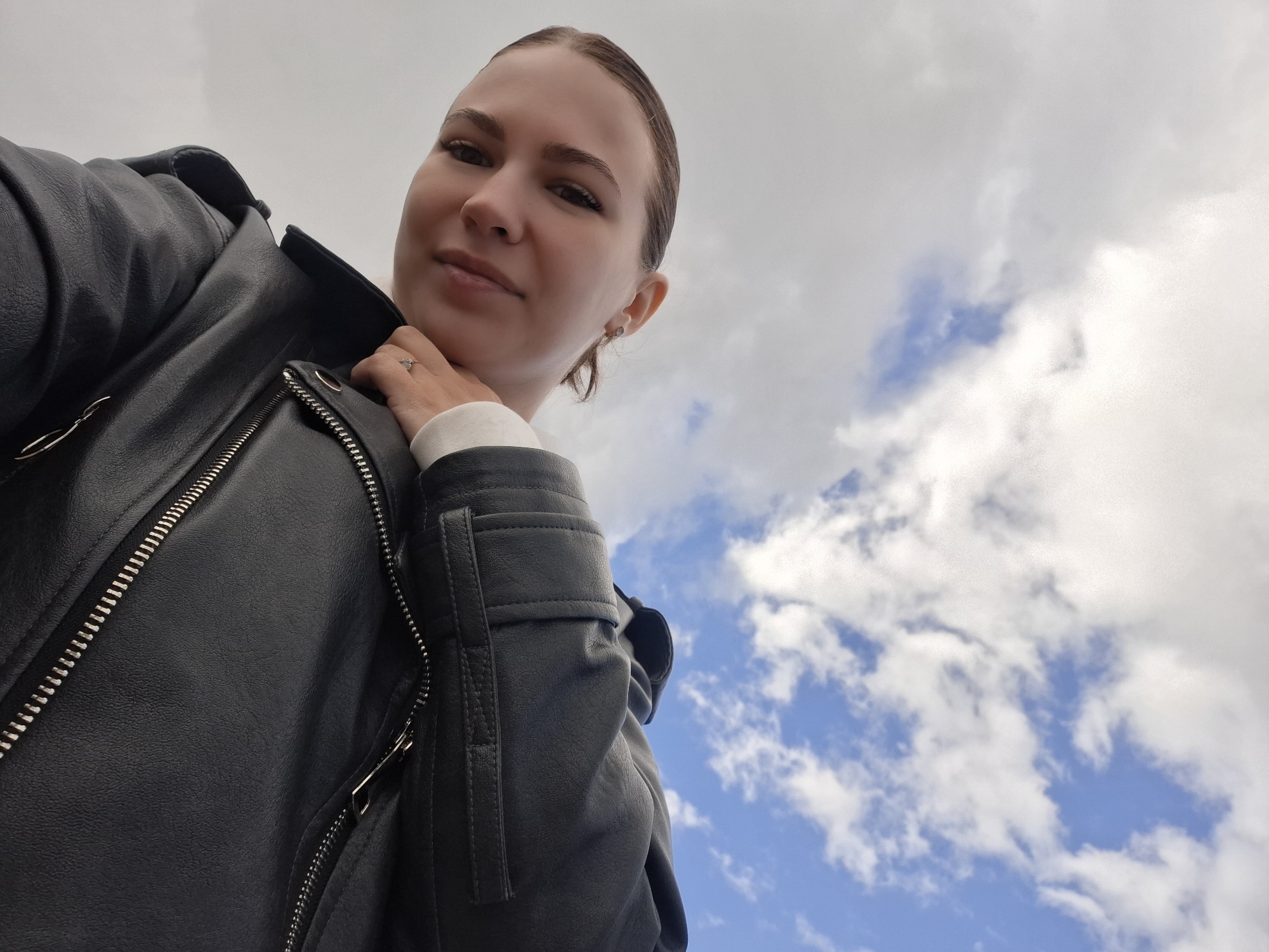 A young woman poses in a low-angle shot against a backdrop of a partly cloudy sky. She wears a black leather jacket over a white garment and has her hair pulled back. Her left hand, adorned with a ring, touches her neck. The background consists of a blue sky dotted with white clouds. The location cannot be determined from the image.