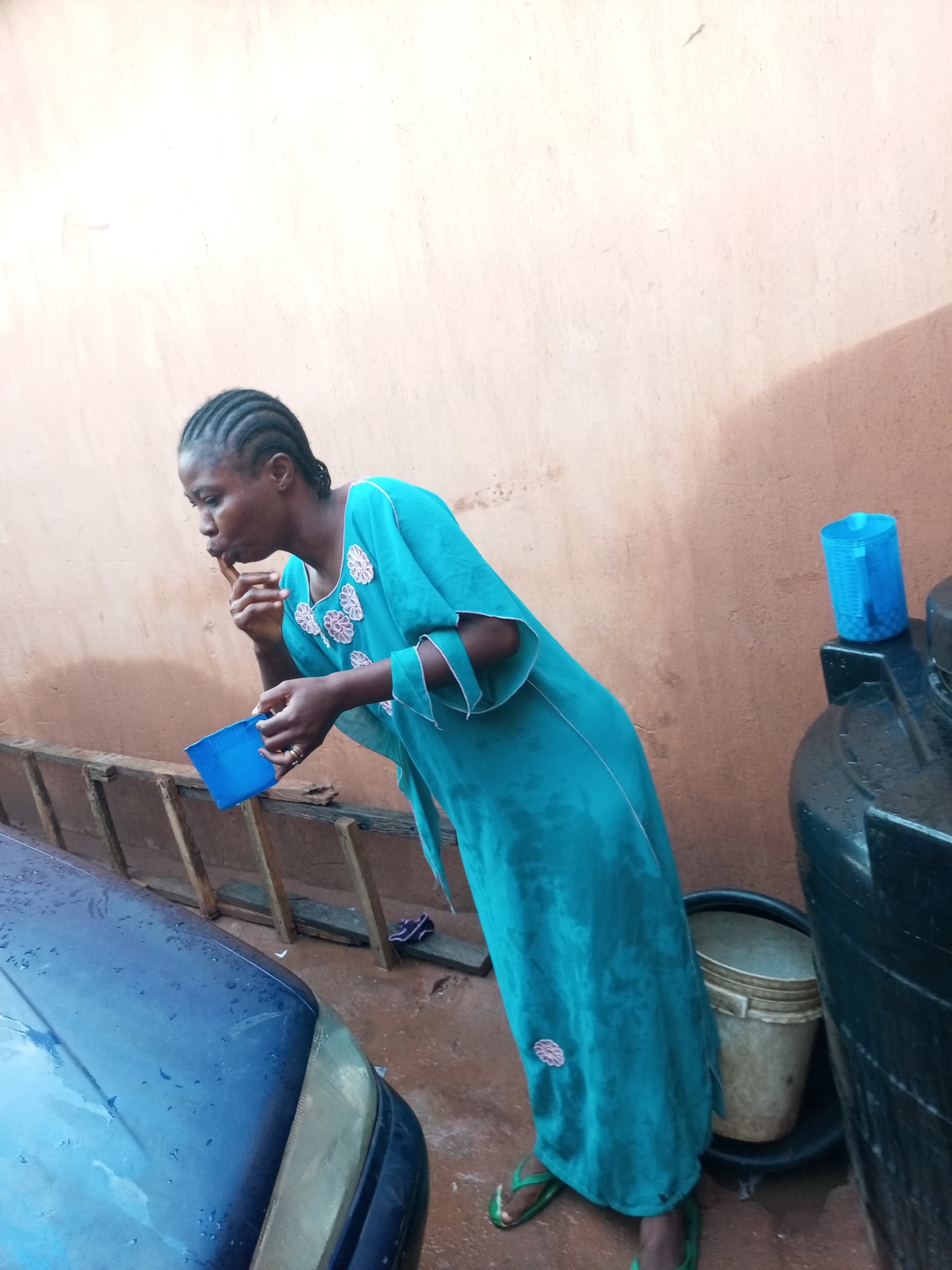 In Tempure, Nigeria, a woman in a teal dress with floral embroidery and green flip-flops is captured outdoors. She is holding a blue plastic cup in her left hand and appears to be bringing her right index finger to her mouth. She is standing next to a blue car, which has water droplets on its surface. Behind her, a wooden structure is visible, and further in the background, a large black water tank with a blue plastic jug on top and a bucket at its base. The ground is wet and appears to be a dirt path. The wall behind the woman is a light brown color.