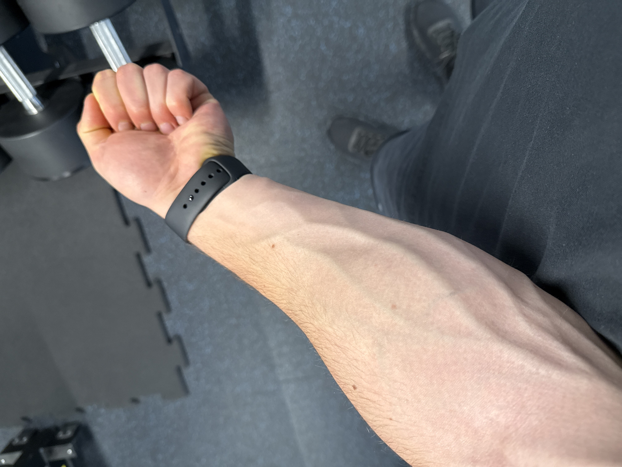 A person's arm is extended towards the camera, showing prominent veins. The person is wearing a black smartwatch. The hand is clenched in a fist, possibly holding a dumbbell. The person is wearing dark pants and grey shoes. The background shows a gym floor with rubber tiles and a dumbbell rack. The location cannot be determined from this image.