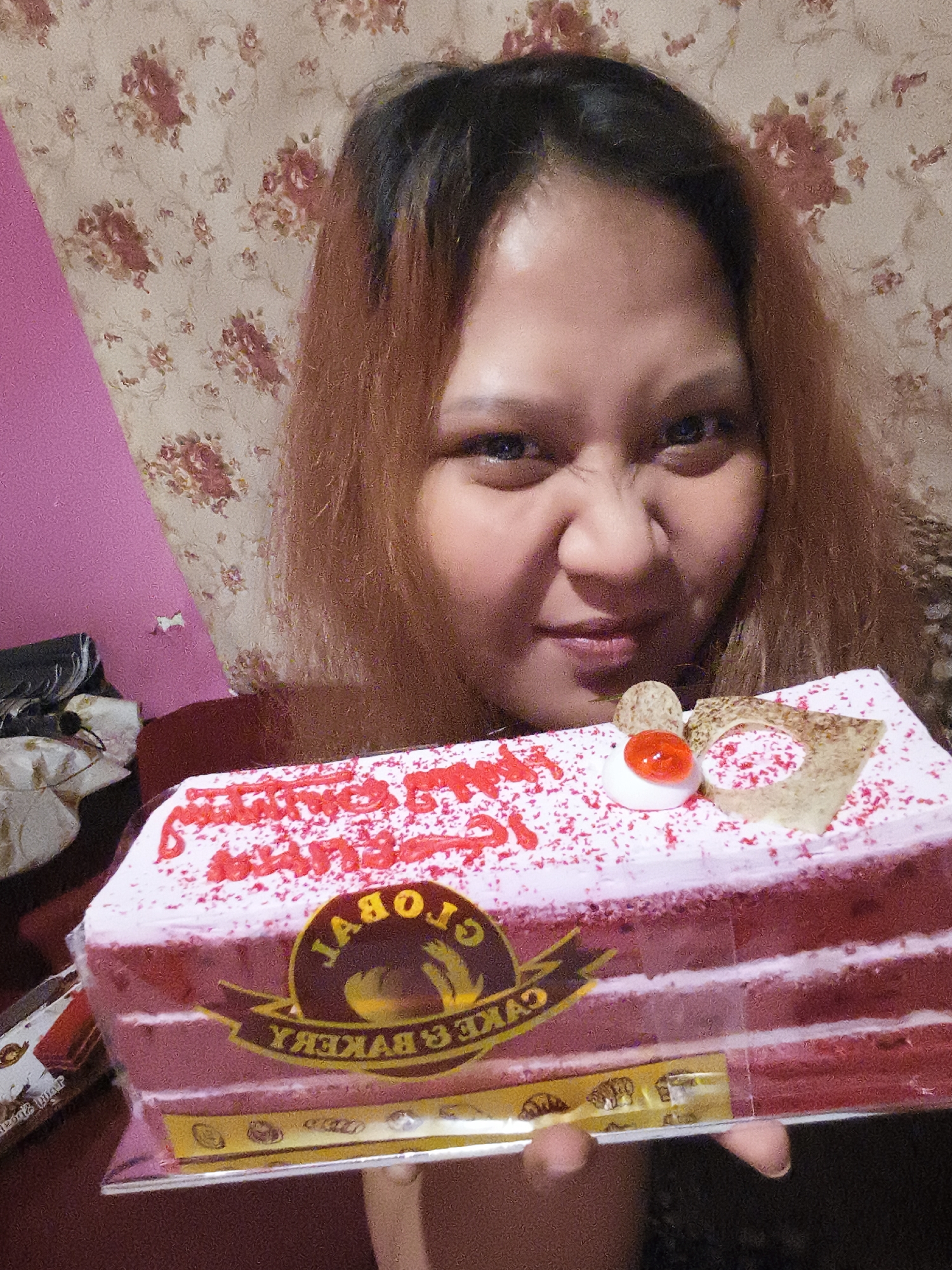 A woman with long brown hair and fair skin is pictured holding a rectangular red velvet cake with white frosting and red sprinkles. The cake is decorated with a small white meringue topped with a red jelly, and a piece of pastry. The woman is making a playful grimace with her eyes squinted. She is wearing a patterned top with dark and light brown colors. Behind her, the wall is covered with a floral wallpaper in shades of beige, pink, and red. To the left of the woman, a pink wall is visible, and further to the left, a black trash bag and some white objects are stacked. The cake has a label that reads "GLOBAL CAKE & BAKERY". The image appears to be taken indoors, possibly in a home setting in the city of Depok, Indonesia.