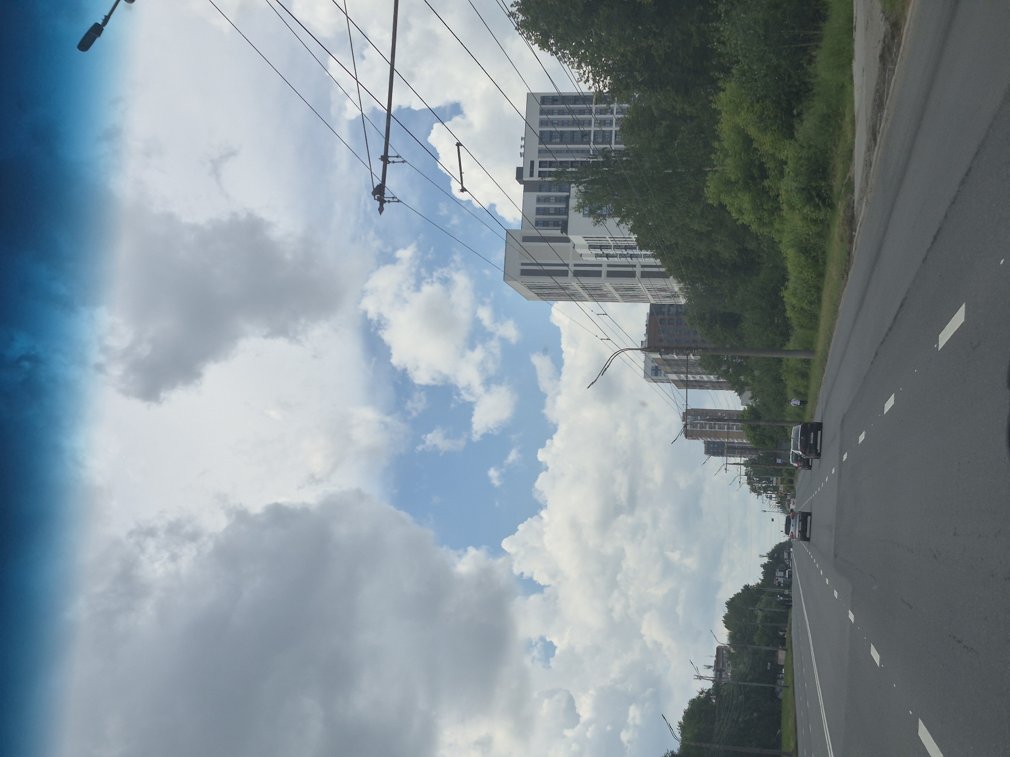The image depicts a multi-lane asphalt road in Izhevsk, Russia, with a backdrop of modern high-rise buildings and lush green trees. The sky is partly cloudy, with a clear blue sky visible between large, white clouds. Power lines crisscross the sky, supported by poles. Several cars are driving on the road, and a lone pedestrian is walking on the sidewalk. The scene appears to be a typical urban landscape with a mix of residential and infrastructure elements. The overall impression is one of a bright, possibly warm, day.