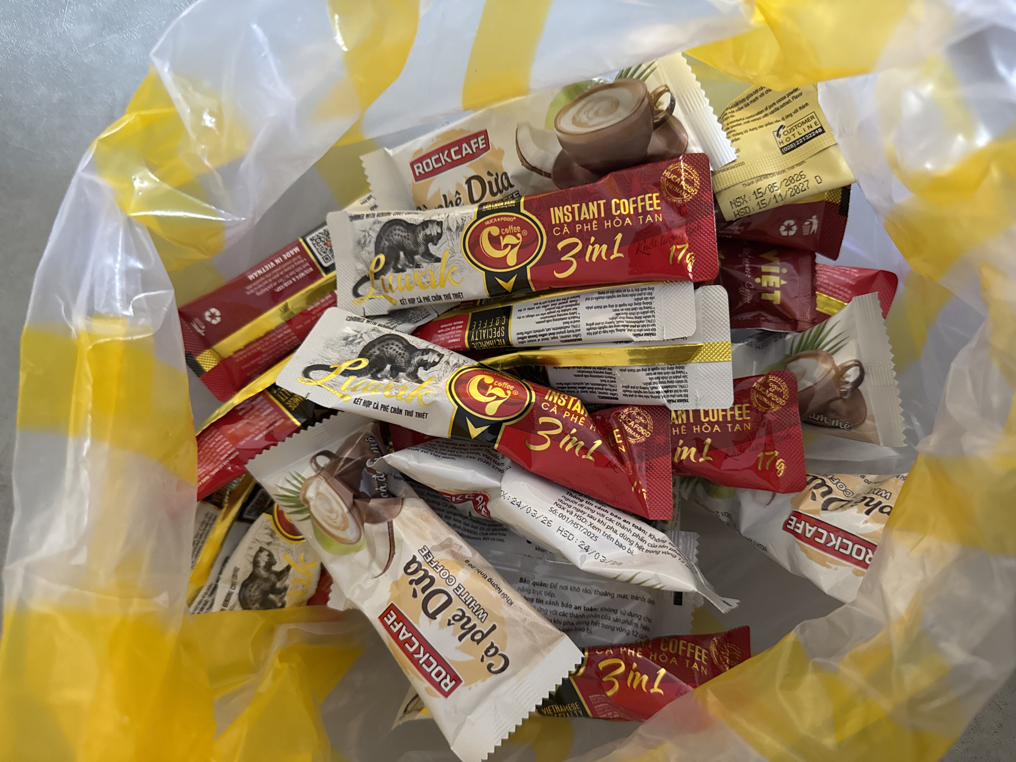 A plastic grocery bag, primarily yellow with white accents, is filled with numerous small, individually packaged coffee sachets. The sachets are a mix of white, red, and yellow, featuring various branding, including "ROCKCAFE," "G coffee," and "INSTANT COFFEE." Some sachets display images of coffee cups, coconuts, or what appear to be civet cats. The bag is positioned outdoors on a textured grey surface, possibly a sidewalk or outdoor table, in a location that is not identifiable as Солонешное, Russia. There are no people or animals present.
