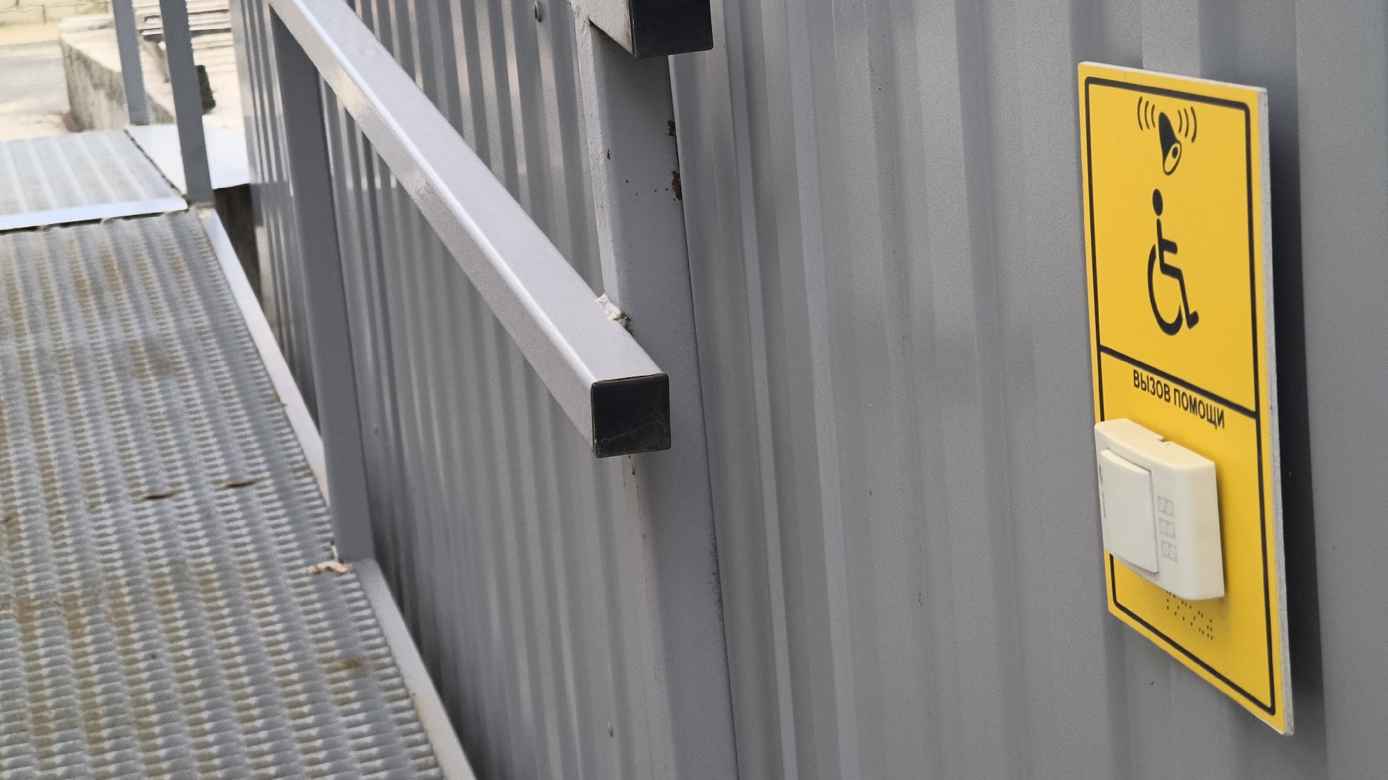 The image shows a yellow sign with a black wheelchair symbol and text in Russian reading "ВЫЗОВ ПОМОЩИ" (call for help). Below the sign is a white electronic call button. This sign and button are mounted on a ribbed metal wall. To the left of the wall is a metal ramp with a grated surface. A metal handrail runs diagonally across the image, attached to the wall. The scene appears to be outdoors, possibly near a public building or facility. There are no people visible in the image. The location is described as Mikhaylovka, Russia.