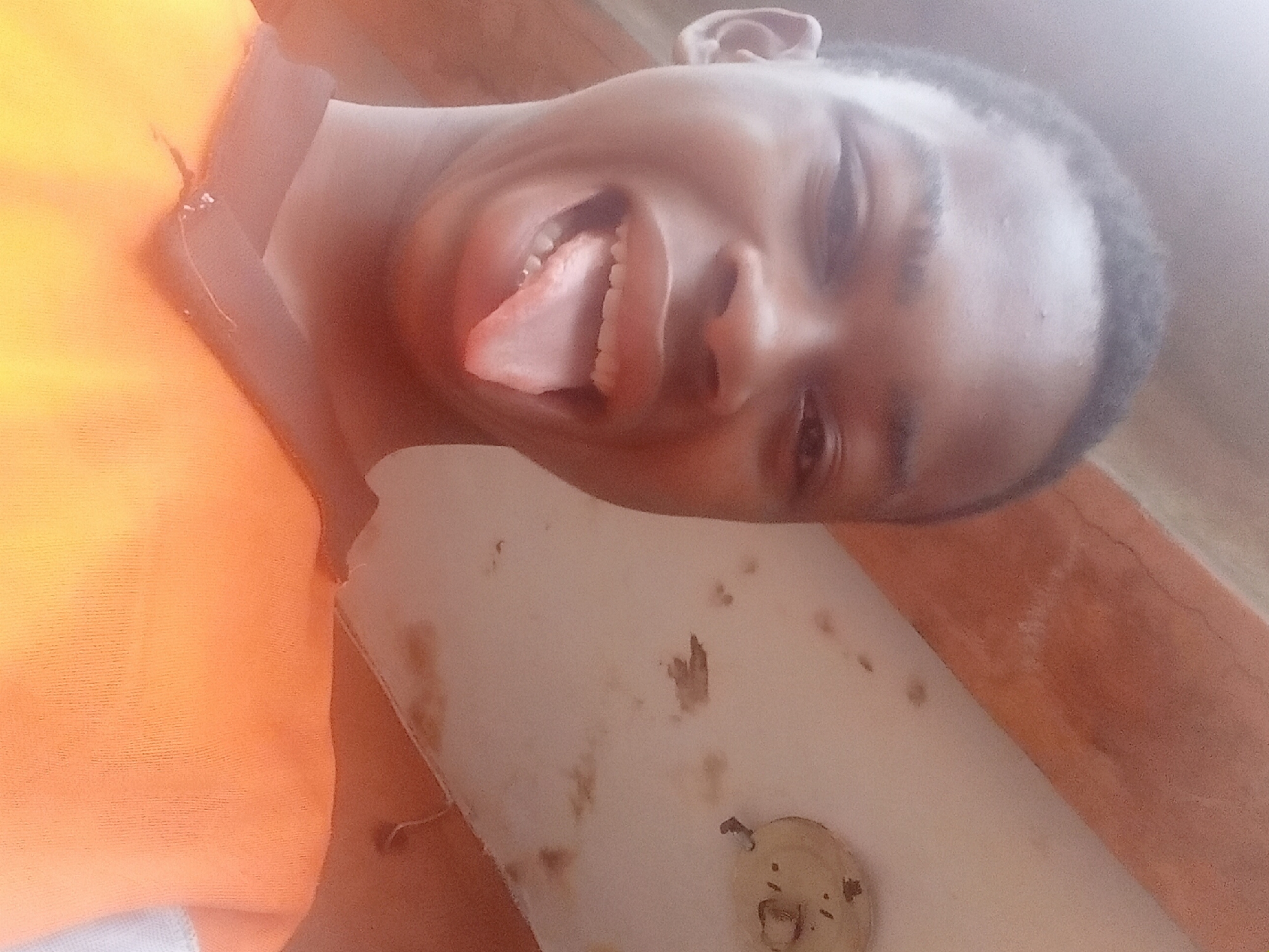 A medium shot angled to the right depicts a young, dark-skinned person with a close haircut, sticking out their tongue. The person is wearing an orange garment on the left side of the frame. The background is a white, possibly stained or dirty, surface with brown discoloration. The location, Nkpor, Nigeria, cannot be verified from the image. No actions or interactions are evident beyond the person sticking out their tongue.