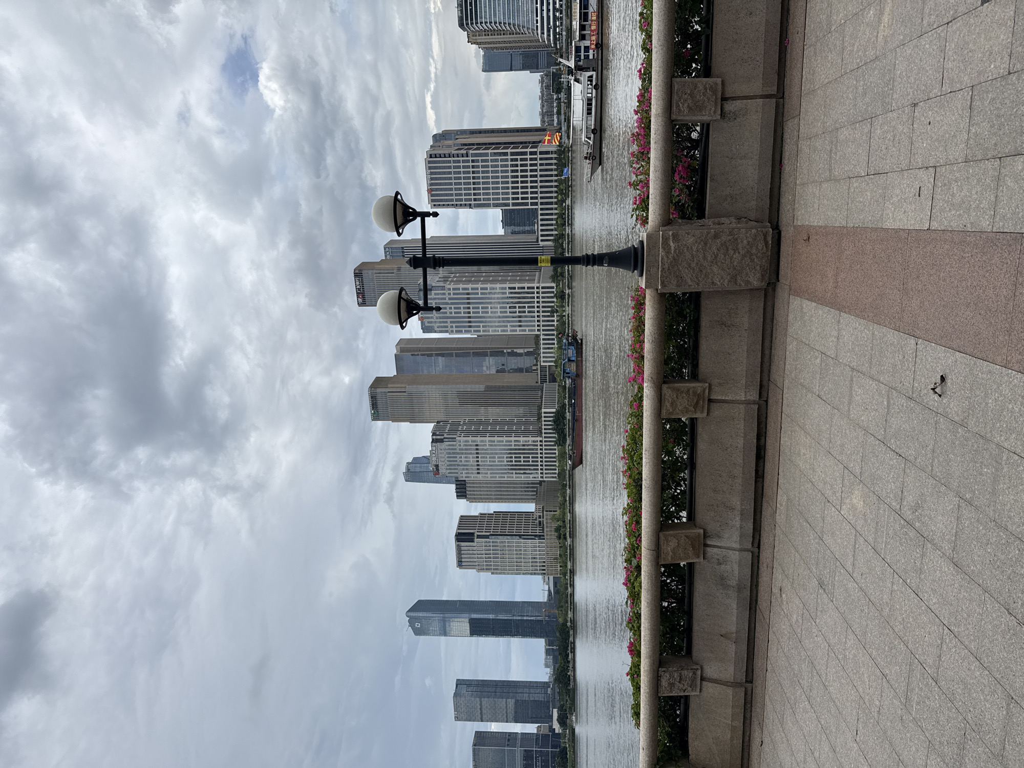 An overcast sky looms over a waterfront cityscape in Haizhu District, China. The foreground features a paved walkway with a low, stone wall adorned with planters of pink flowers. A lamppost with two globes stands prominently on the right side of the walkway. Beyond the wall, a wide river separates the promenade from a dense cluster of modern skyscrapers that dominate the skyline. Several boats, including a large red vessel and a white passenger boat, navigate the river. The architecture of the buildings is varied, with sleek glass facades and more textured stone exteriors. The overall scene captures a blend of urban development and the natural element of the river.