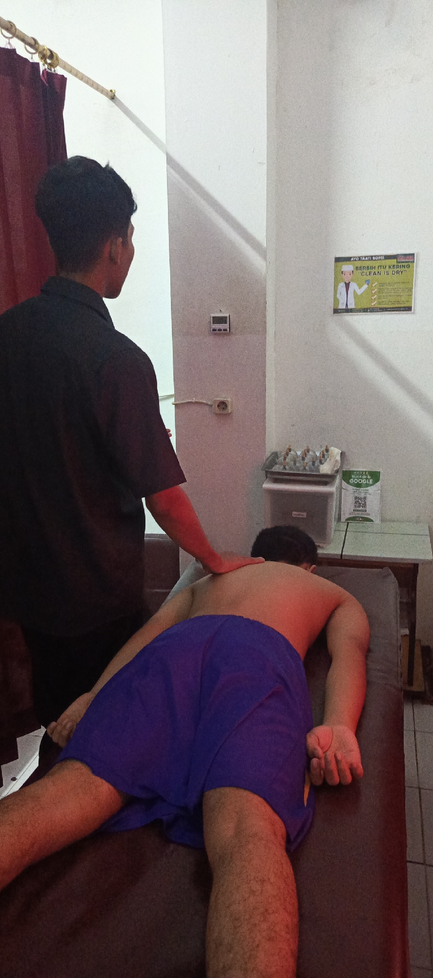 In the city of Sukaresmi, Indonesia, a masseuse, dressed in a black collared shirt, is performing a back massage on a client lying face down on a dark brown massage table. The client is wearing blue shorts. The scene appears to be indoors, with a maroon curtain visible on the left side of the image. The wall behind the subjects is light-colored and features a digital clock, an electrical outlet, and a framed poster with text in Indonesian and English. To the right, a white shelf holds several small bottles, and a green sign with a QR code is also present. Red lighting is cast upon the client's back and legs.