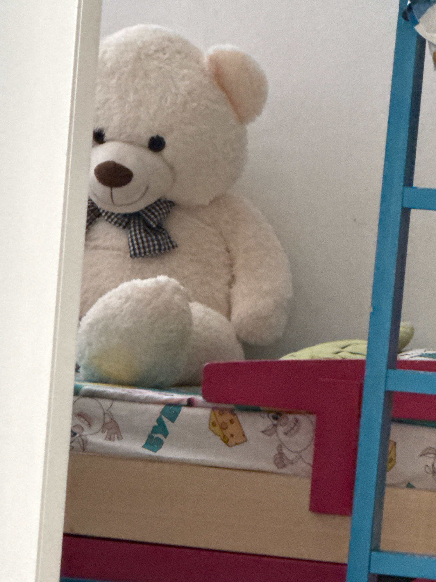 A white teddy bear with a blue and white checkered bow is sitting on a bed. The bed has a light beige mattress and a bedspread with cartoon mice and cheese slices. A pink rail separates the bed from a blue ladder. The scene appears to be in a bedroom. The location in Netanya, Israel is not discernible from the image.