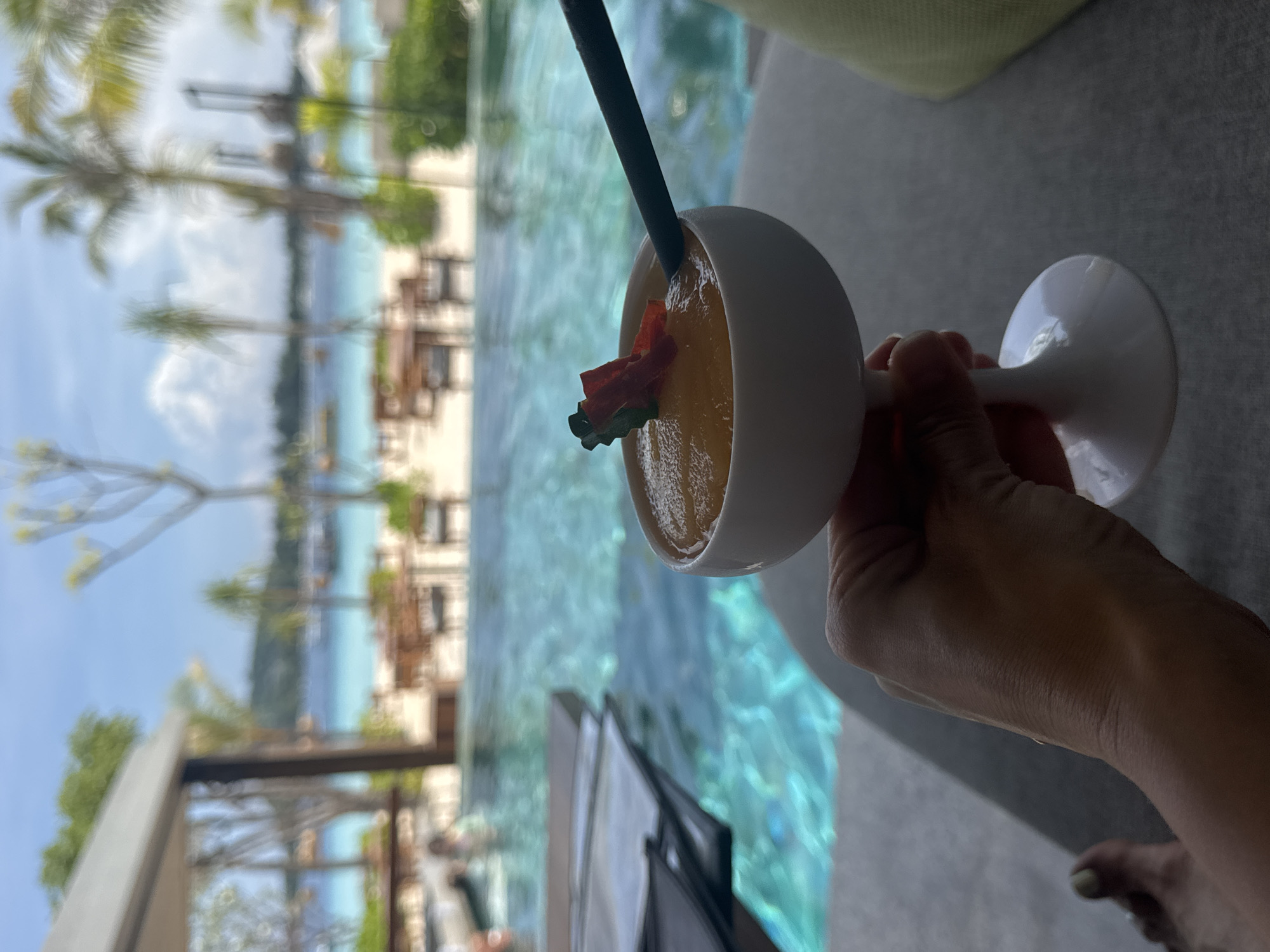 The scene presents an individual holding a white stem glass filled with a vibrant orange liquid, presumably a cocktail, garnished with a piece of red fruit and a dark straw. The backdrop reveals a swimming pool with turquoise water, beyond which lies a beach with palm trees and various structures, suggesting a resort or tropical setting. The individual's hand is visible holding the stem glass. The image is taken in Unknown, Indonesia.