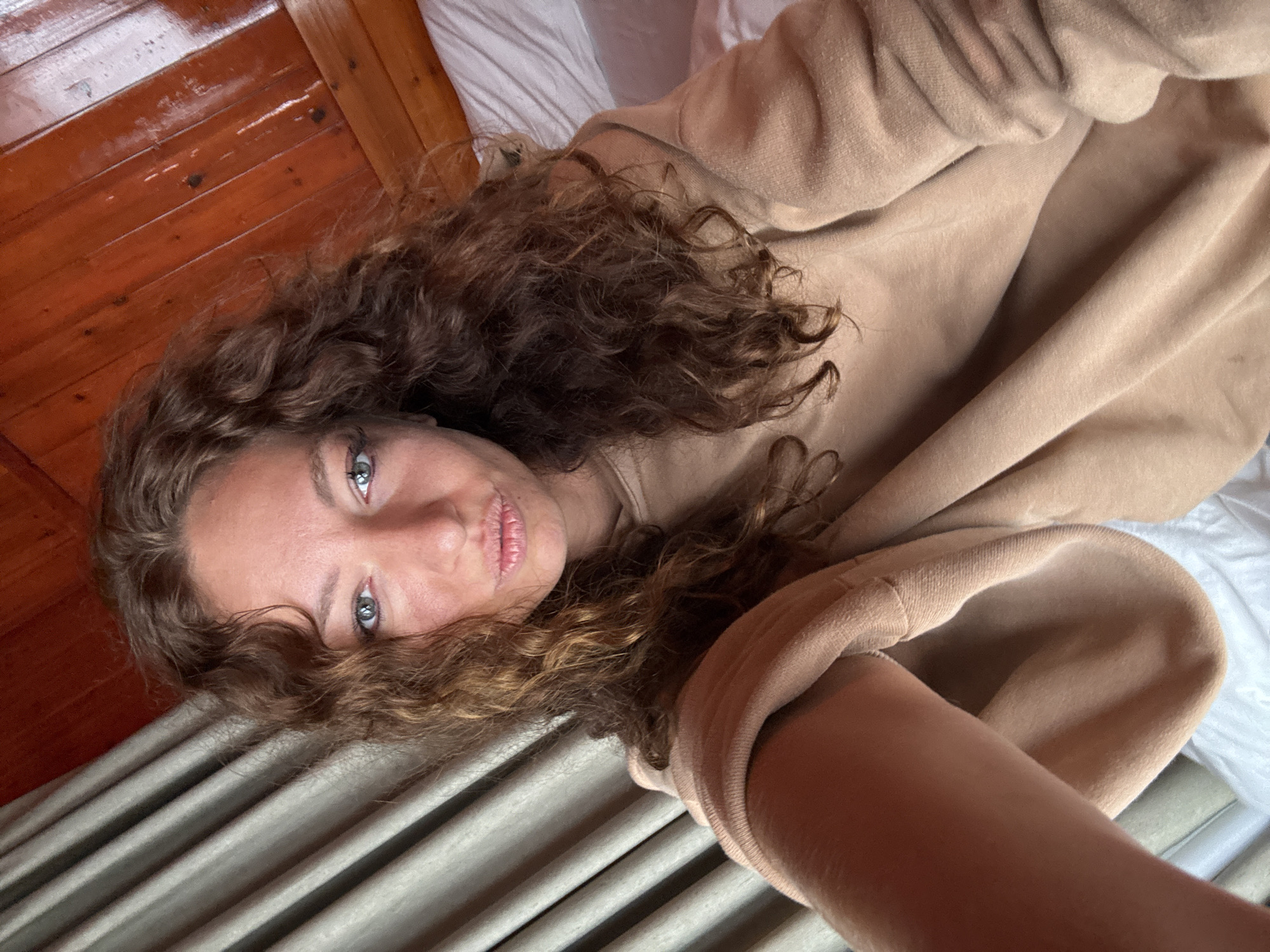 A young woman lies on a bed in a room with wooden walls and a gray and white patterned bedspread. She is wearing a tan sweatshirt, and her curly brown hair is spread out around her head. Her eyes are light-colored. She appears to be taking a selfie. Given the prompt, the location is presumed to be Binh Lu, Vietnam, but there are no visual cues to confirm this location.