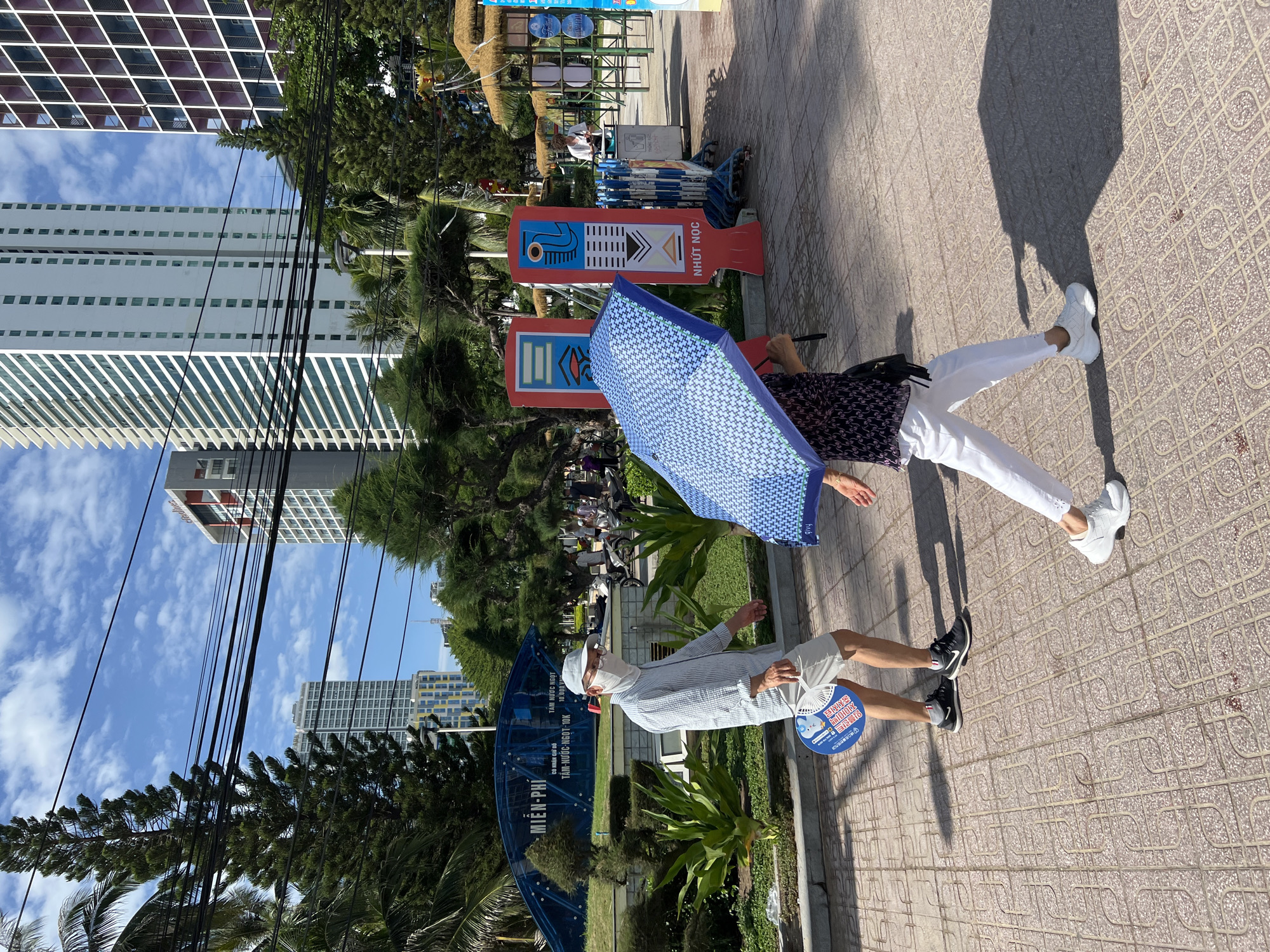 The image depicts a street scene in Nha Trang, Vietnam, with a focus on two pedestrians. On the left, a person wearing a light blue, pinstriped long-sleeved shirt, light-colored shorts, a white mask, and a light blue cap walks to the right. They are holding a white fan with blue text and graphics in their left hand and have a dark blue and white circular object, possibly a cooler bag or a round umbrella base, on the ground to their left. Their right leg is slightly bent, and they are wearing black athletic shoes. To their right and slightly ahead, another person, mostly obscured by a blue and white patterned umbrella, walks to the right. This person wears a dark patterned top and white pants with frayed edges at the ankles. They have on white athletic shoes. The ground is paved with square tiles arranged in a grid pattern. In the background, tall buildings are visible, some with many windows. Lush green trees and plants are scattered throughout the scene. A blue arched structure with text on it is visible behind the first pedestrian. Other people and parked motorcycles are in the distance, and a sidewalk with stalls covered by thatched roofs can be seen further back on the right. The sky is blue with scattered white clouds.