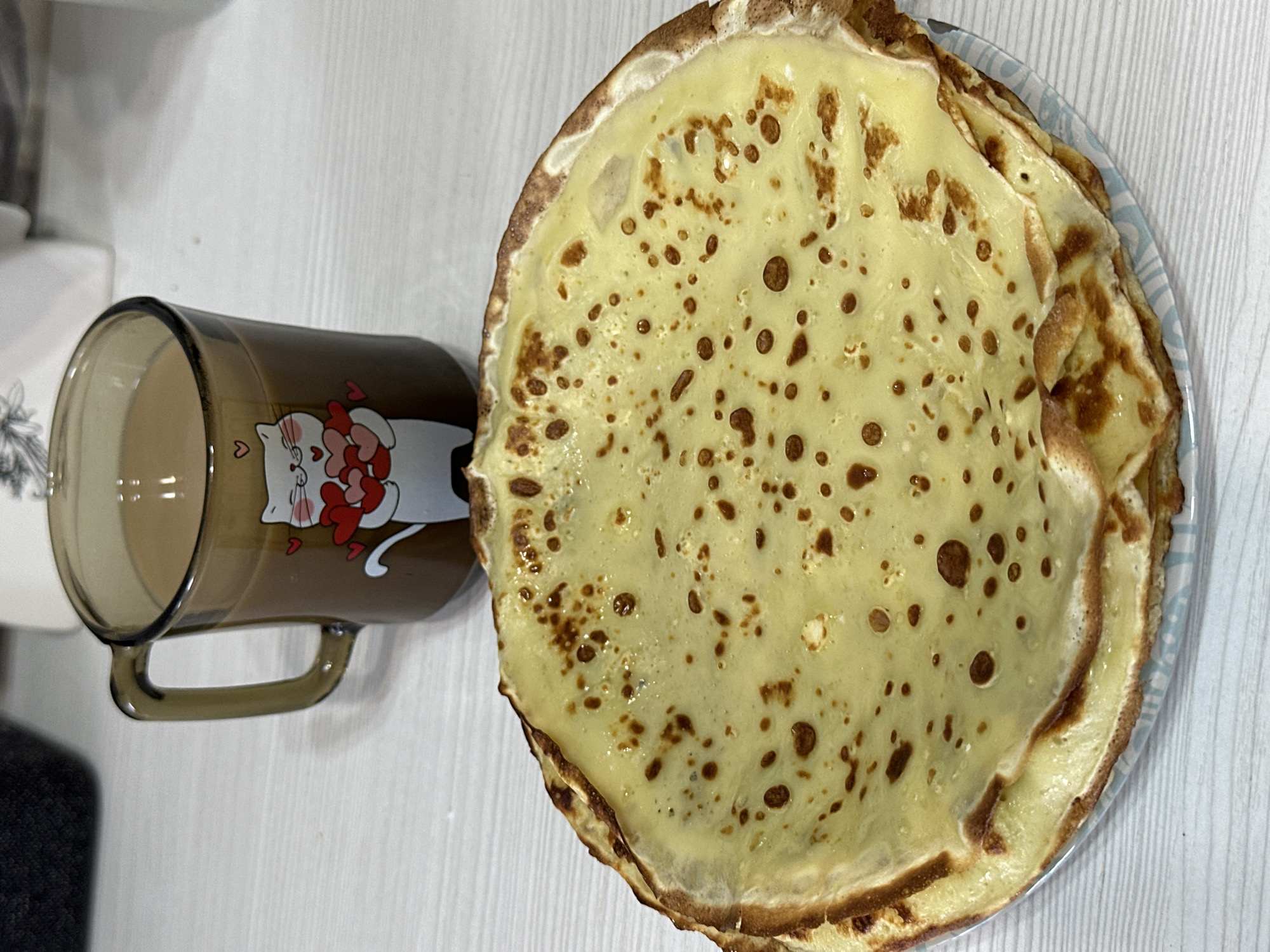 The image, reportedly from the city of Chaikovsky, Russia, presents an indoor still life scene. In the foreground, a stack of multiple thin, round pancakes or crepes, predominantly light yellow with scattered darker brown cooking spots, rests on a white plate featuring a subtle blue patterned rim. The top pancake exhibits a slight sheen. Behind and slightly to the left of the pancakes, a translucent amber-tinted glass mug with a handle holds a light brown liquid, filled approximately two-thirds. The mug is decorated with a white cartoon cat holding several red hearts, accompanied by smaller floating red heart motifs. All objects are positioned on a light-colored table surface exhibiting fine horizontal graining. The background is out of focus, showing indistinct darker and lighter elements. No individuals are visible.