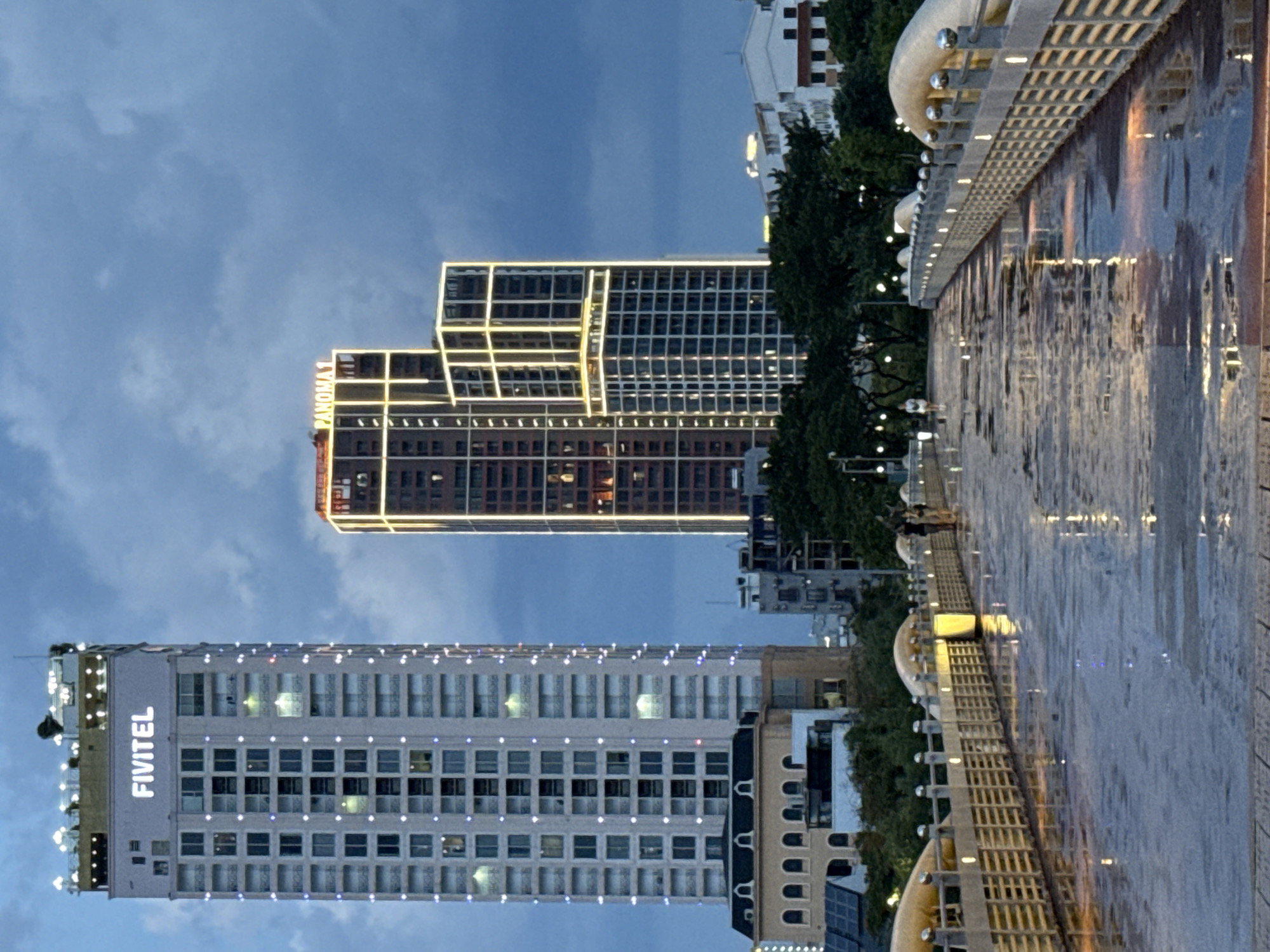 This media file depicts an urban scene at dusk in Đà Nẵng, Vietnam. The perspective is tilted, suggesting it might have been taken from a bridge or elevated walkway overlooking a body of water.
The primary subjects are two prominent skyscrapers and a water feature. The skyscraper on the left is modern and white, adorned with numerous small lights that create a festive or decorative effect. The text "FIVITEL" is clearly visible in large, illuminated letters on its side, identifying it as a hotel or telecommunications company building. To its right, another skyscraper is visible, partially obscured by trees. This building features a grid-like pattern of yellow lights outlining its structure, giving it a modern, illuminated aesthetic. The text "PANOMA" is visible on this building as well.
The foreground is dominated by a wide, wet surface, likely a promenade or walkway beside a river or canal. The surface is reflective, mirroring the lights from the surrounding buildings and the sky, creating a shimmering effect. A decorative railing with a unique curved design and small spherical embellishments runs along the edge of this walkway. Lights are also visible along the edge of the walkway, further illuminating the scene.
The sky is a deep blue, characteristic of twilight, with some clouds present. The overall atmosphere is serene and possibly slightly humid, given the wet surfaces. The time of day is clearly evening or dusk, as artificial lights are prominent and the natural light is fading. No people are clearly discernible in the image, but the presence of a walkway suggests it's a public space. The overall impression is one of a vibrant, modern cityscape at night, with a focus on architectural lighting and reflections.
