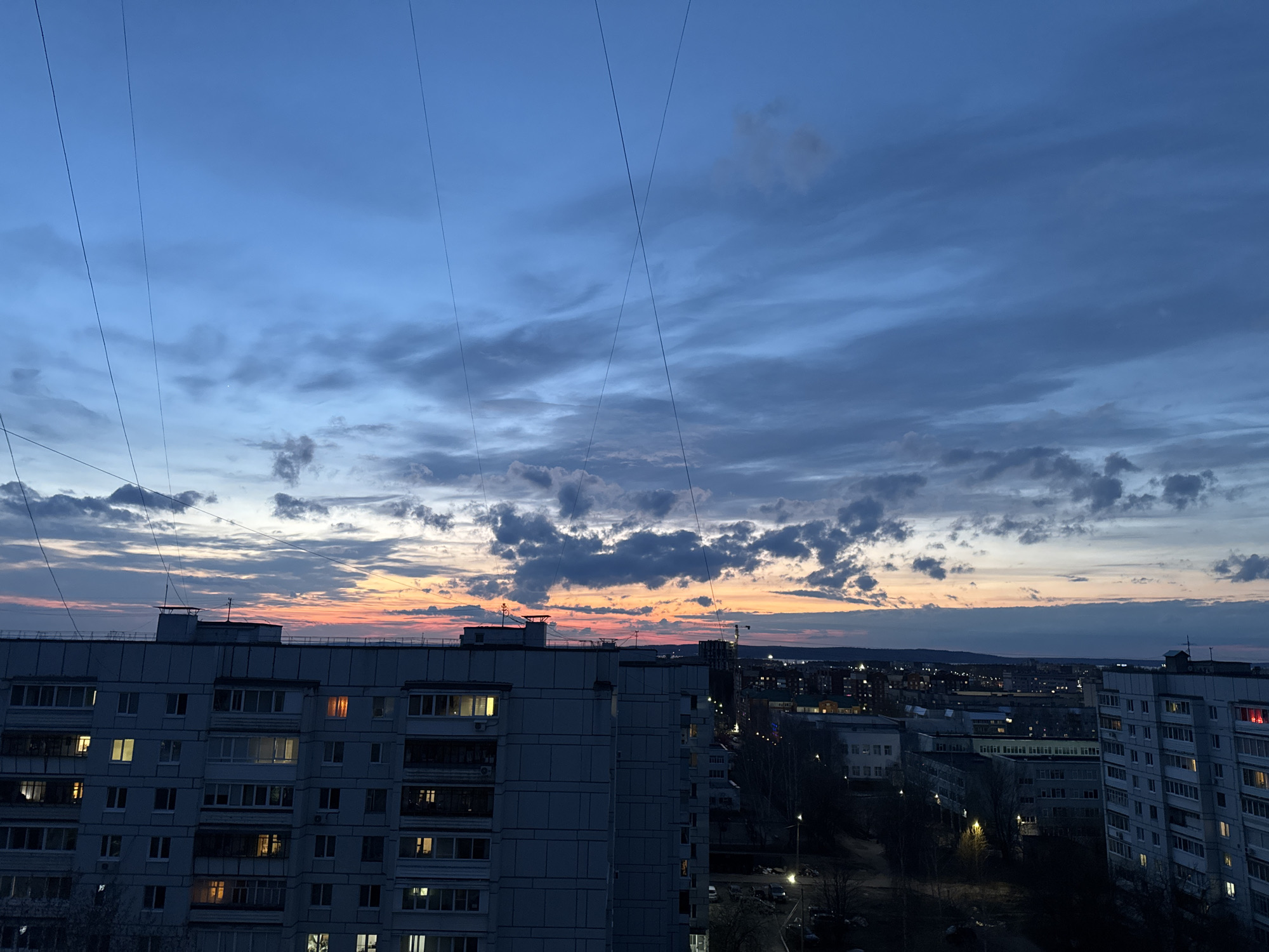 This media file captures an urban landscape at twilight, viewed from a high vantage point, likely a window, in Chaikovsky, Russia. The scene depicts the transition from day to night, or vice versa, with a dramatic sky dominating the upper portion of the image.
**Subjects:** The primary subjects include a large, multi-story residential building (apartment block) occupying the lower left and central foreground. Its facade is a light gray, with numerous windows, several of which are illuminated with warm, yellow light, indicating human activity inside. In the background, other residential and perhaps commercial buildings are visible, forming a dense cityscape that stretches towards the horizon. City lights, street lamps, and car lights dot the darker background. Overhead, multiple thin, dark lines, likely wires or cables, crisscross the sky, adding a foreground element.
**Activity or event taking place:** The image portrays a peaceful evening or early morning in a city. The illuminated windows suggest residents are either winding down for the night or beginning their day. The overall activity is the quiet progression of time in an urban environment.
**Notable details:**
* **Time of day:** The vivid colors on the horizon suggest either a recent sunset or an impending sunrise. A band of warm orange and pink hues transitions into cool blues and grays higher in the sky.
* **Weather:** The sky is partly cloudy, with scattered cumulus clouds reflecting the changing light. The weather appears calm, with no signs of precipitation.
* **Location cues:** The architecture of the buildings is typical of urban residential areas, particularly in Eastern Europe. The provided context specifies the location as Chaikovsky, Russia.
* **Emotions:** The scene evokes a sense of tranquility, urban familiarity, and the quiet beauty of twilight.
* **Visible text:** There is no discernible text within the image.