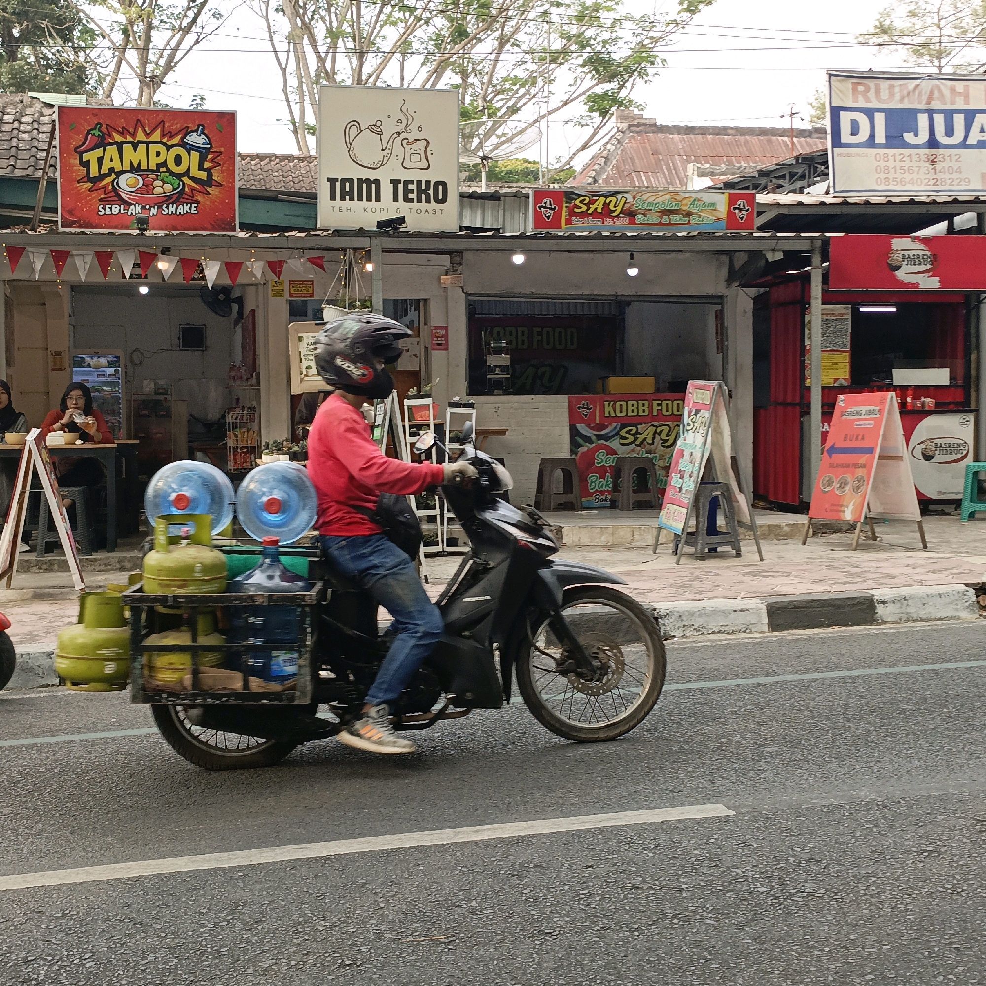 A man in a red long-sleeved shirt and blue jeans rides a black motorcycle with a rack carrying two green propane tanks and two large blue-lidded water jugs. The motorcycle is on a paved road in the city of Salatiga, Indonesia. In the background, there are several food stalls with signs. One sign, prominently displayed, reads "TAMPOL SEBLAK 'N SHAKE". Another sign says "TAM TEKO TEH, KOPI & TOAST". Further down the street, a sign for "RUMAH DI JU..." (likely "Rumah Dijual" meaning "House for Sale") is visible. A woman in a hijab is seated at a table outside one of the stalls, eating. The sky is overcast, and trees with bare branches are visible above the buildings. The scene appears to be a street-level view of a commercial area.