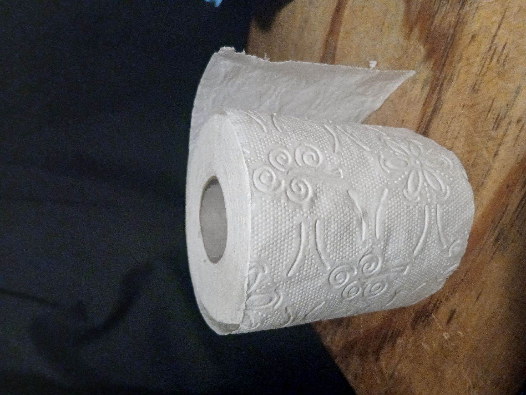 A roll of white toilet paper with a floral embossed pattern is positioned on a wooden surface against a dark, out-of-focus background. The toilet paper roll is slightly unrolled, revealing a torn edge of paper. The wooden surface appears to be a tabletop or plank, showing grain and some wear. The lighting is dim, casting subtle shadows. There are no people present in the image. The scene does not provide any specific information or context to place it within the city of Douai, France.
