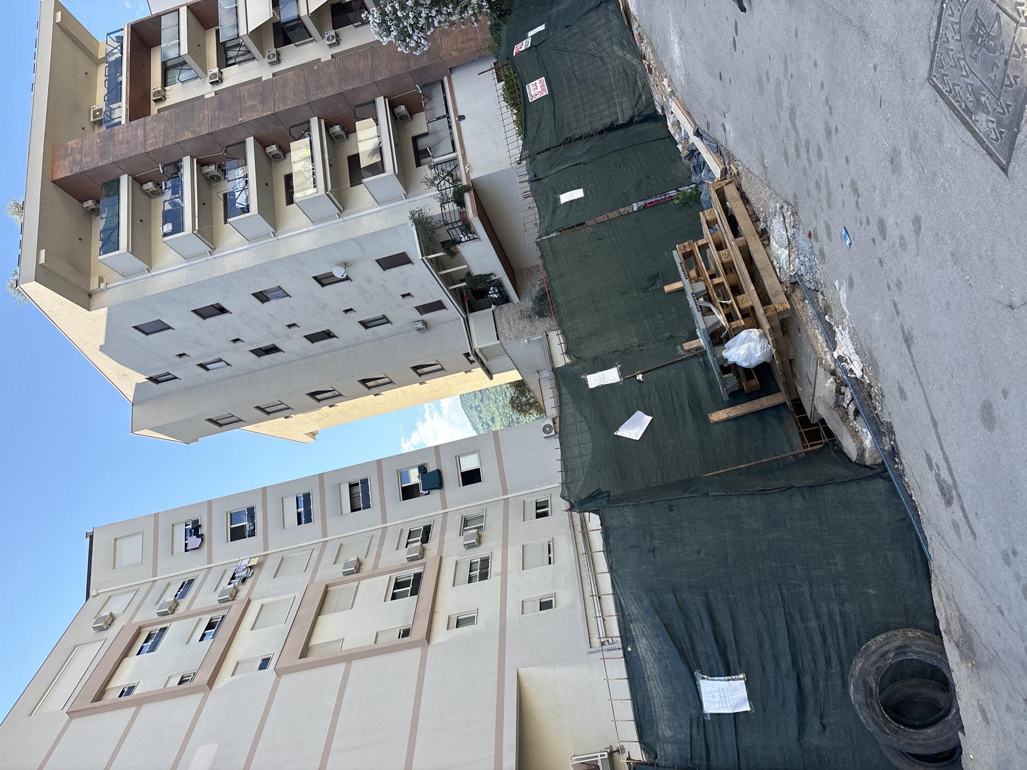 The image shows a construction site in the city of Budva, Montenegro, as evidenced by the Serbian or Montenegrin text on a sign attached to the green mesh fencing, which reads "ULAZ ZABRANJEN" (Entrance Forbidden). Two modern apartment buildings stand tall in the background, with balconies and windows. The foreground is dominated by a green construction fence enclosing the site, with various construction materials and debris visible, including wooden pallets, tires, and a white plastic bag. The scene suggests an ongoing construction or renovation project, with the fence serving to secure the area and prevent access. The overall impression is one of urban development and activity.