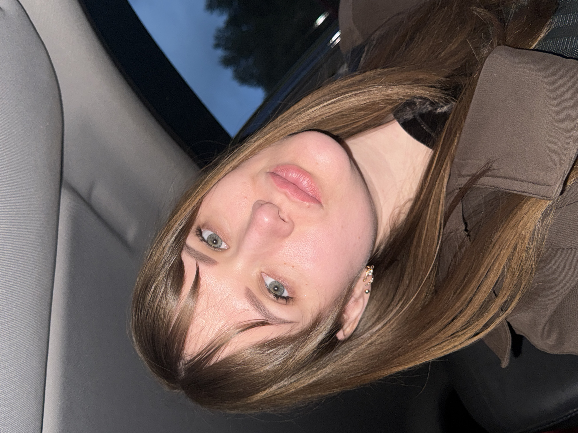 A woman with light skin, green eyes, and light brown hair is pictured in an indoor setting, possibly the interior of a car, as indicated by the presence of a car seat and window frame. The woman is wearing a brown garment, possibly a jacket. Her ear is adorned with multiple piercings and jewelry. The background is blurred, obscuring details of the outdoor environment visible through the window. The location in the city of Chaikovsky, Russia is not discernible from the image.