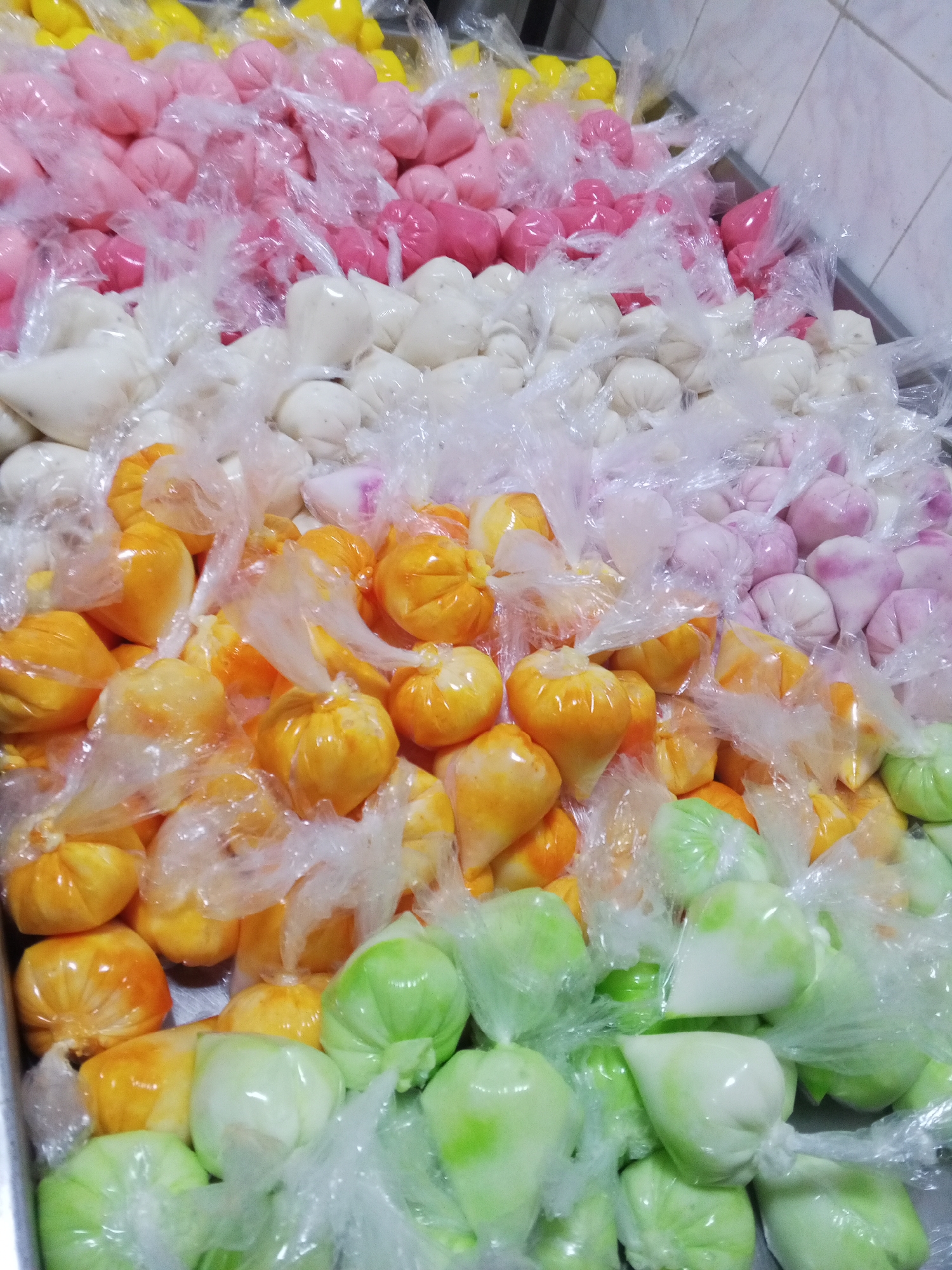 The image shows a stainless steel tray filled with small, colorful, globular-shaped items individually wrapped in clear plastic bags. The colors of the items include yellow, pink, white, orange, light purple, and green. The items appear to be arranged in rows, with each row displaying a single color. The photo was taken in Tinaquillo, Venezuela.