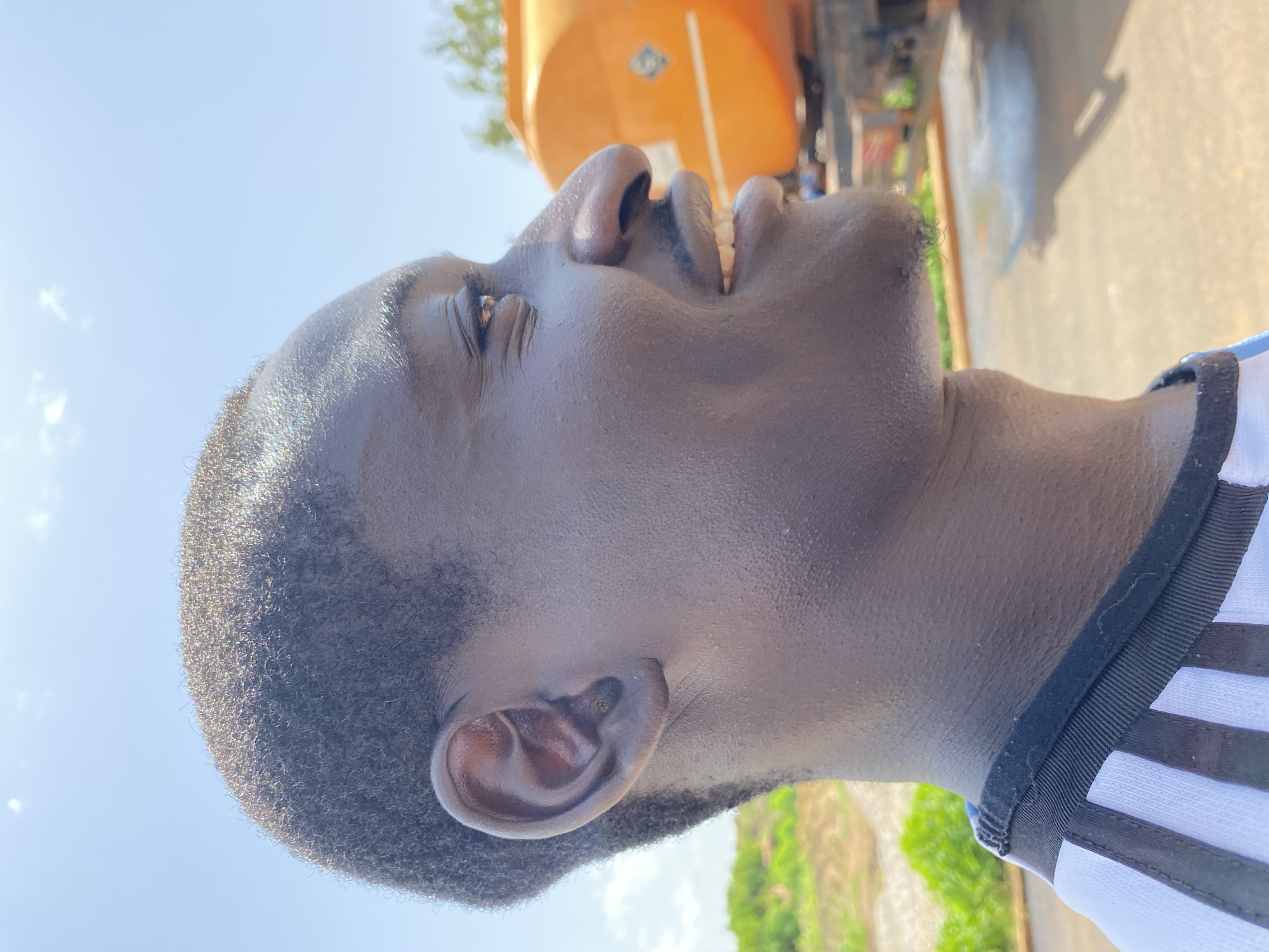 The image is a close-up, profile view of a young Black man's face, looking to the right. He has short, dark hair, dark eyes, and a wide smile revealing his teeth. He is wearing a white shirt with black stripes on the collar. In the background, an orange cylindrical tank truck is visible, along with a blurred landscape of dry earth and green foliage under a bright blue sky. The scene appears to be outdoors, possibly in a dusty or industrial area. There are no explicit indications of the specific location within Bukuru, Nigeria.