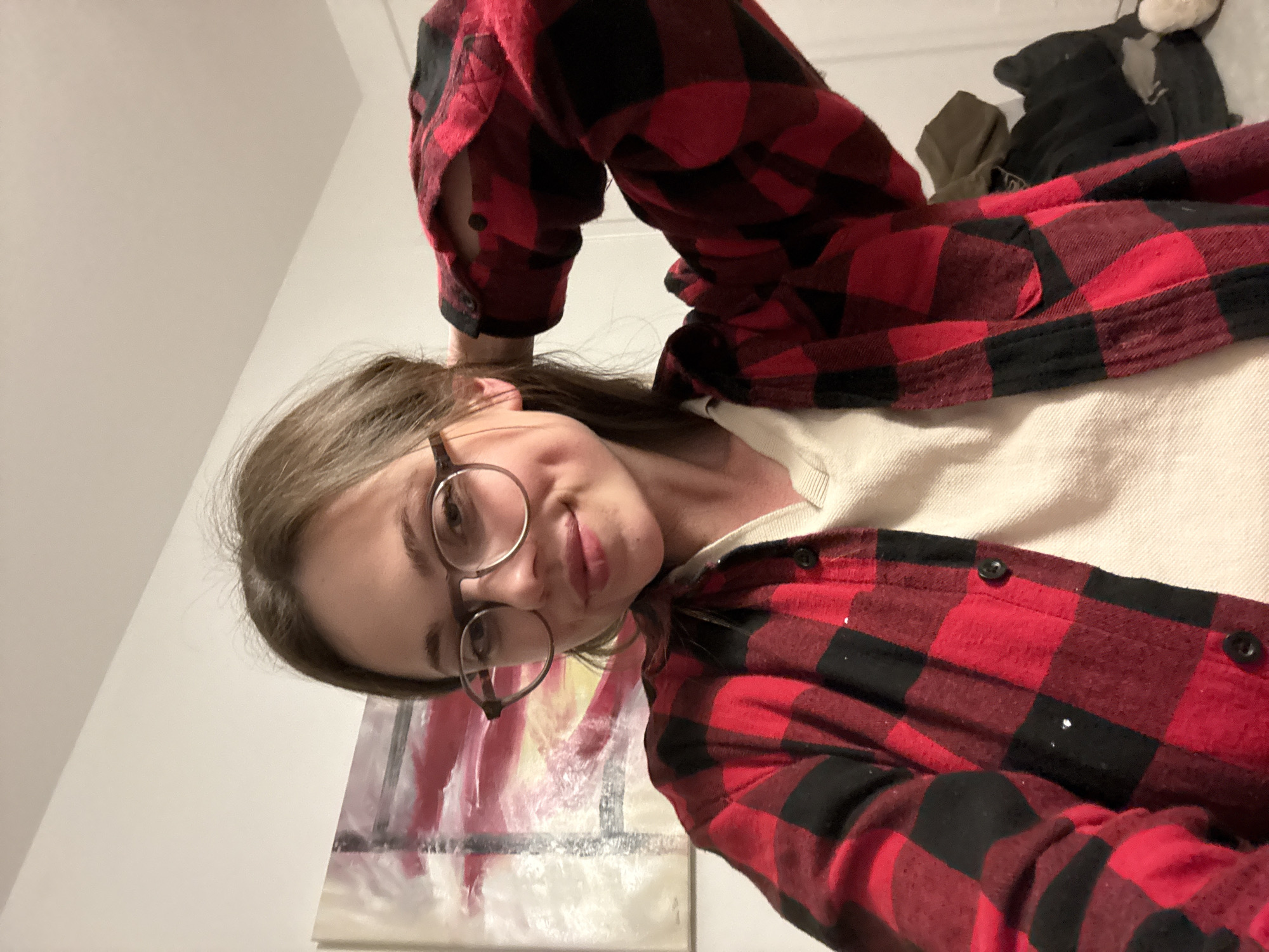 A selfie of a young woman wearing round glasses and a red and black plaid flannel shirt over a white shirt. The background includes a white wall, a piece of art, and some clothing on what appears to be a bed. The location cannot be determined from this image.