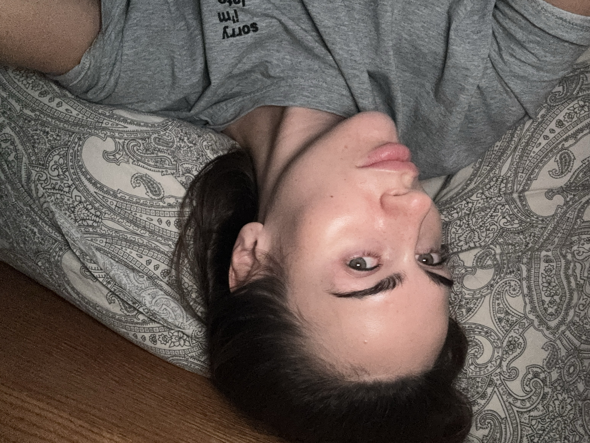 A woman with fair skin and dark hair lies on a patterned pillow, wearing a gray T-shirt with text that reads "Sorry I'm late". Her head is tilted sideways, and she looks towards the camera with gray eyes. The background includes a wooden floor. There is no information regarding the location in the city of Warsaw, Poland.