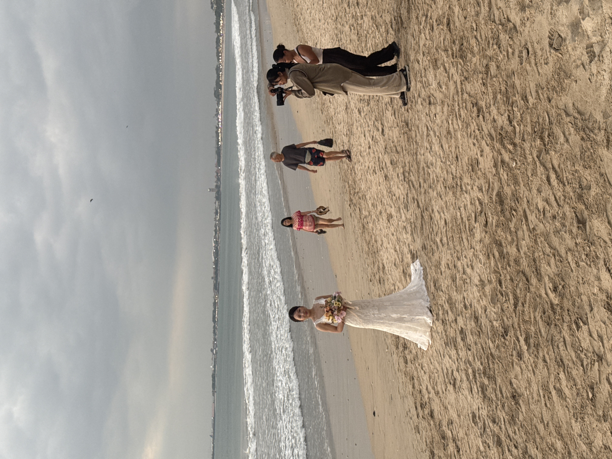 On a sandy beach in Kuta Selatan, Indonesia, a woman in a white wedding dress stands holding a bouquet of flowers. Two women stand to her right, one holding a camera as if photographing her. In the background, a man and another woman walk along the shore, their backs to the camera. The ocean waves crash onto the beach under a cloudy sky, with a distant cityscape visible on the horizon.