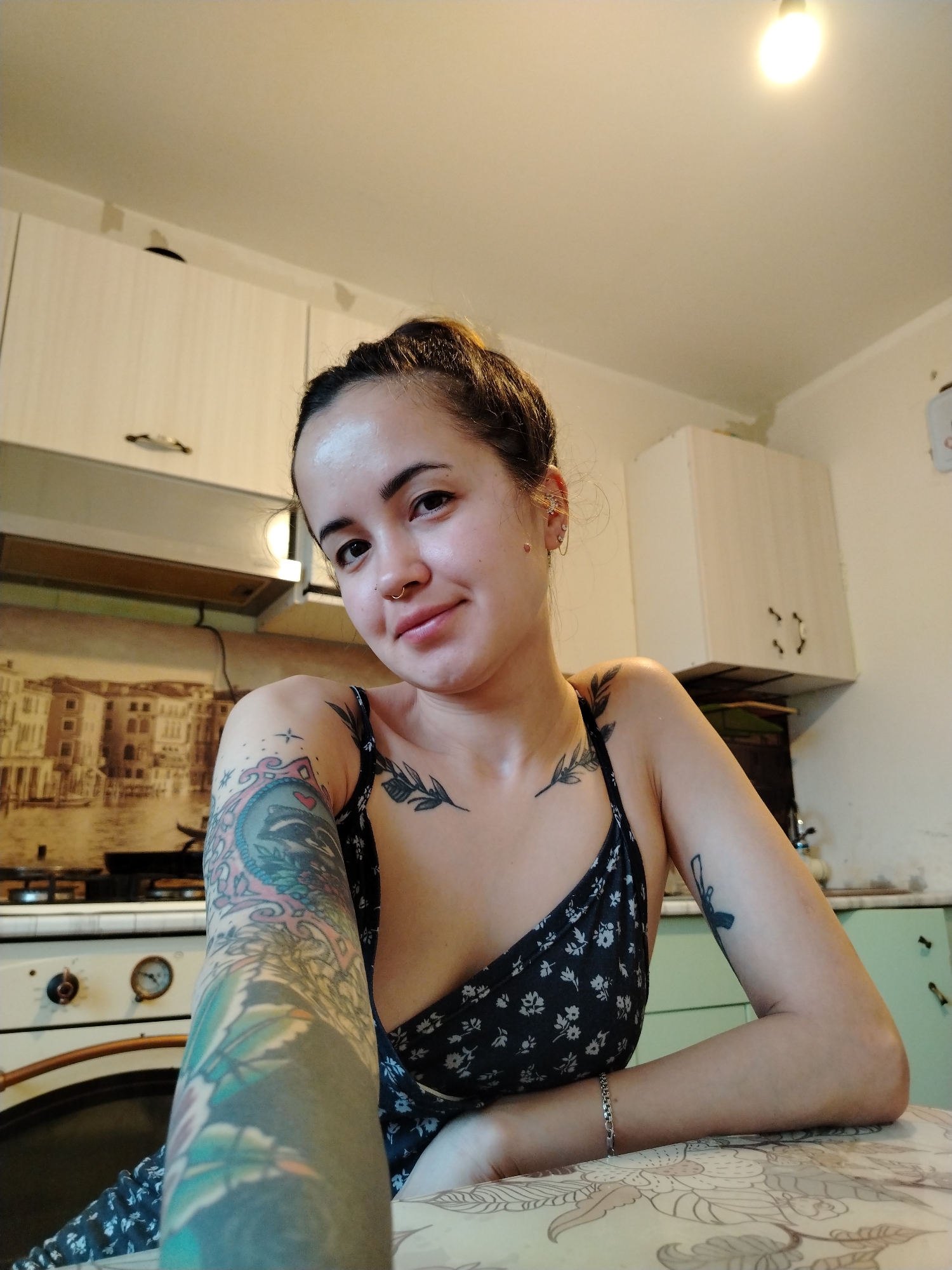 A young woman is seated at a table indoors, likely in a kitchen. She has her hair pulled back in a bun. She is wearing a dark-colored floral print dress and has multiple tattoos visible on her arms, chest, and shoulders. She has a nose ring and multiple piercings in her ear. In the background are kitchen cabinets and an oven. A picture depicting a city reminiscent of Venice, Italy, is behind the oven. A light fixture is visible on the ceiling. The location in the city of Noviy, Russia is not determinable from the visual content.